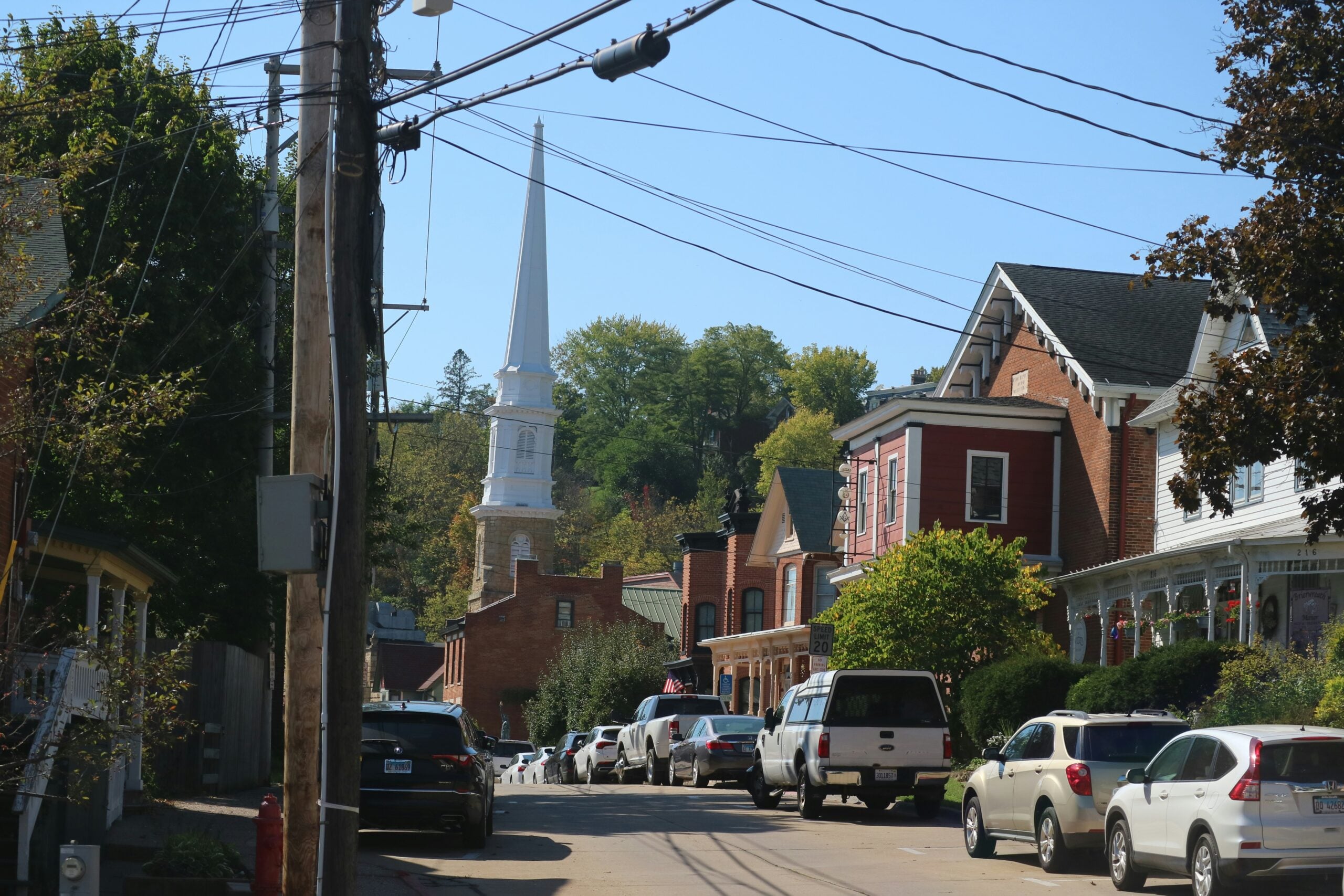 A small town street lined with parked cars and brick houses, with a white church steeple rising in the background under a clear blue sky. Power lines cross overhead and trees are visible behind the buildings.