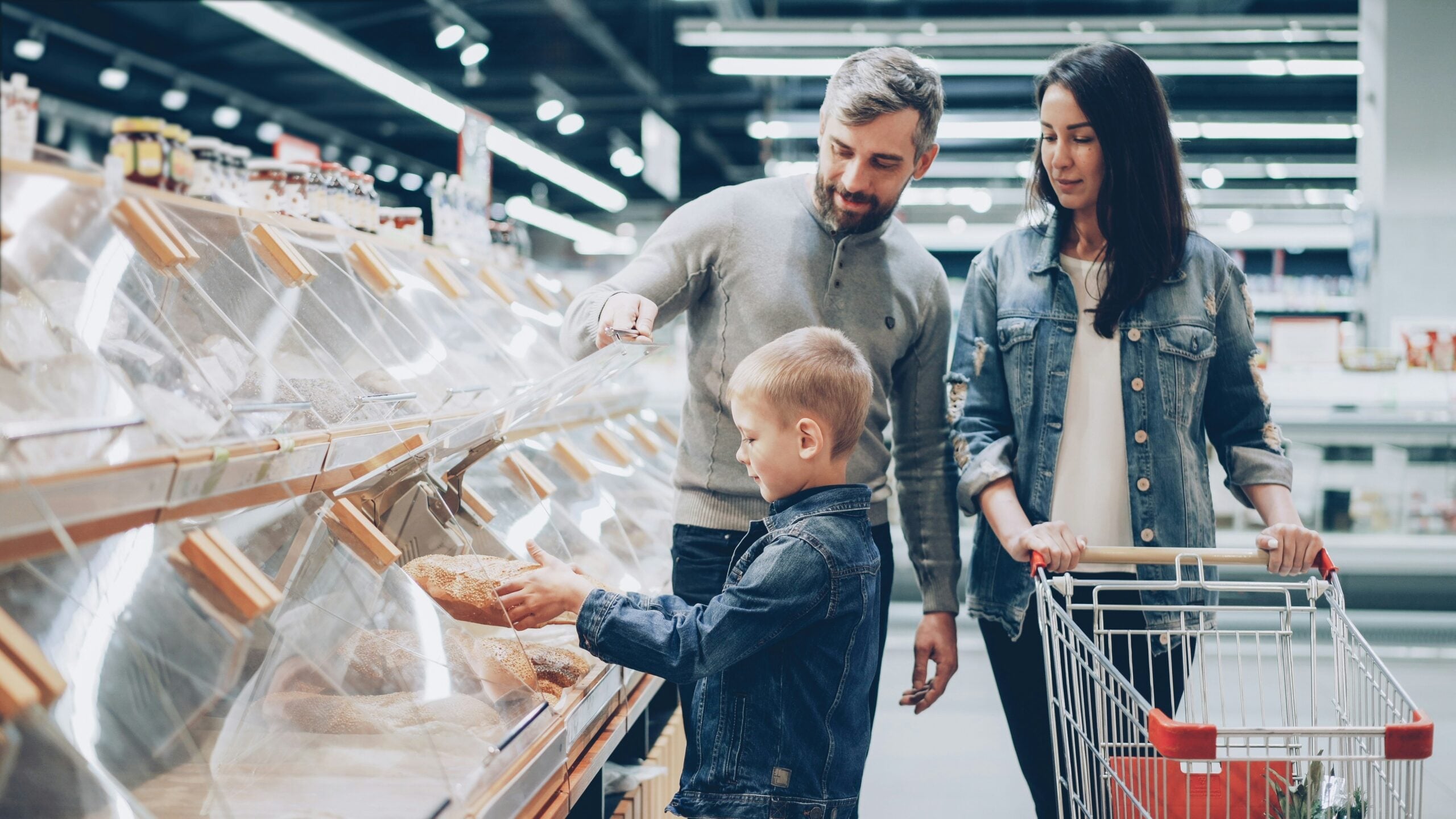 A family of three, two adults and a child, shop for bread in a supermarket. The man and child pick a loaf from a display case while the woman stands nearby with a shopping cart.