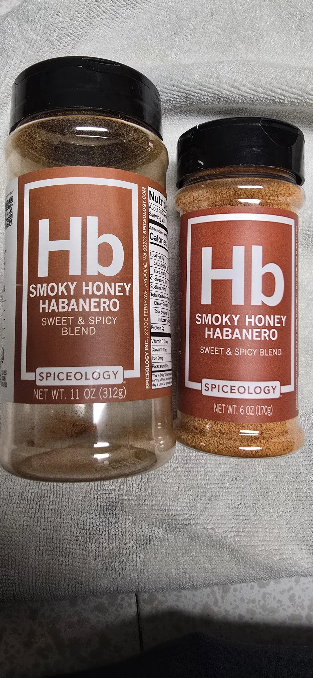Two spice jars labeled "Hb Smoky Honey Habanero Sweet & Spicy Blend" by Spiceology are placed on a light gray textured surface. One jar is 11 oz (312g) and the other is 6 oz (170g).
