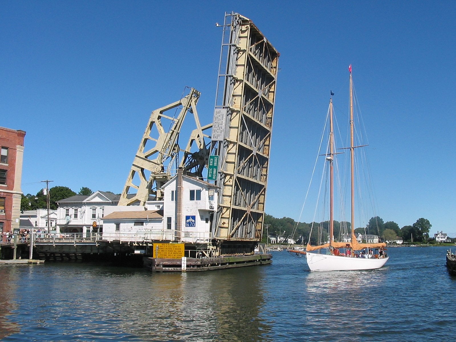 A drawbridge is raised over a river in one of the unique American towns, allowing a white sailboat with tall masts to pass through. Buildings and trees are visible in the background under a clear blue sky.
