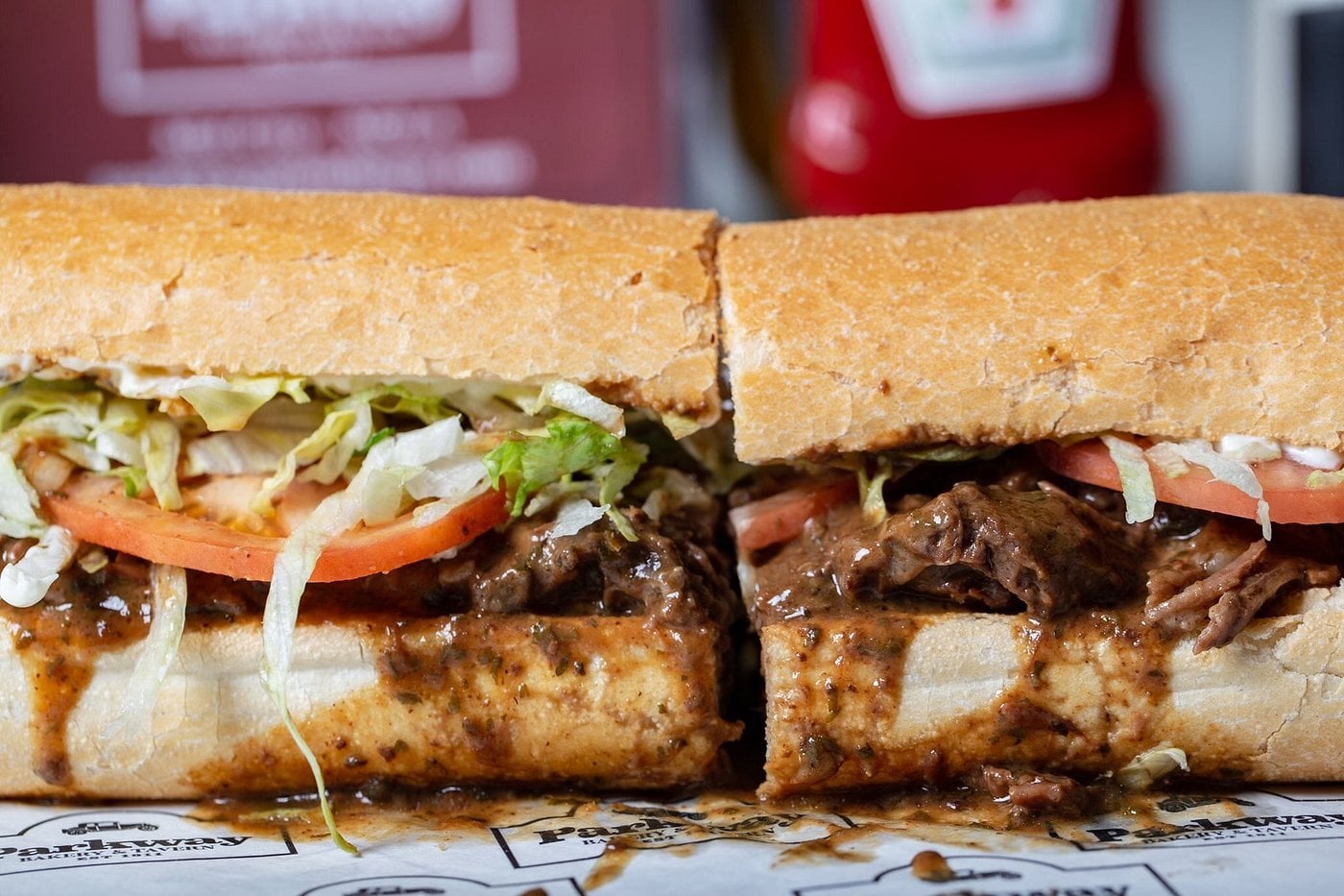 A close-up of a sandwich cut in half, filled with shredded beef in brown gravy, lettuce, tomato, and onions on a hoagie roll—perfect for road trip food stops—with ketchup bottles blurred in the background.