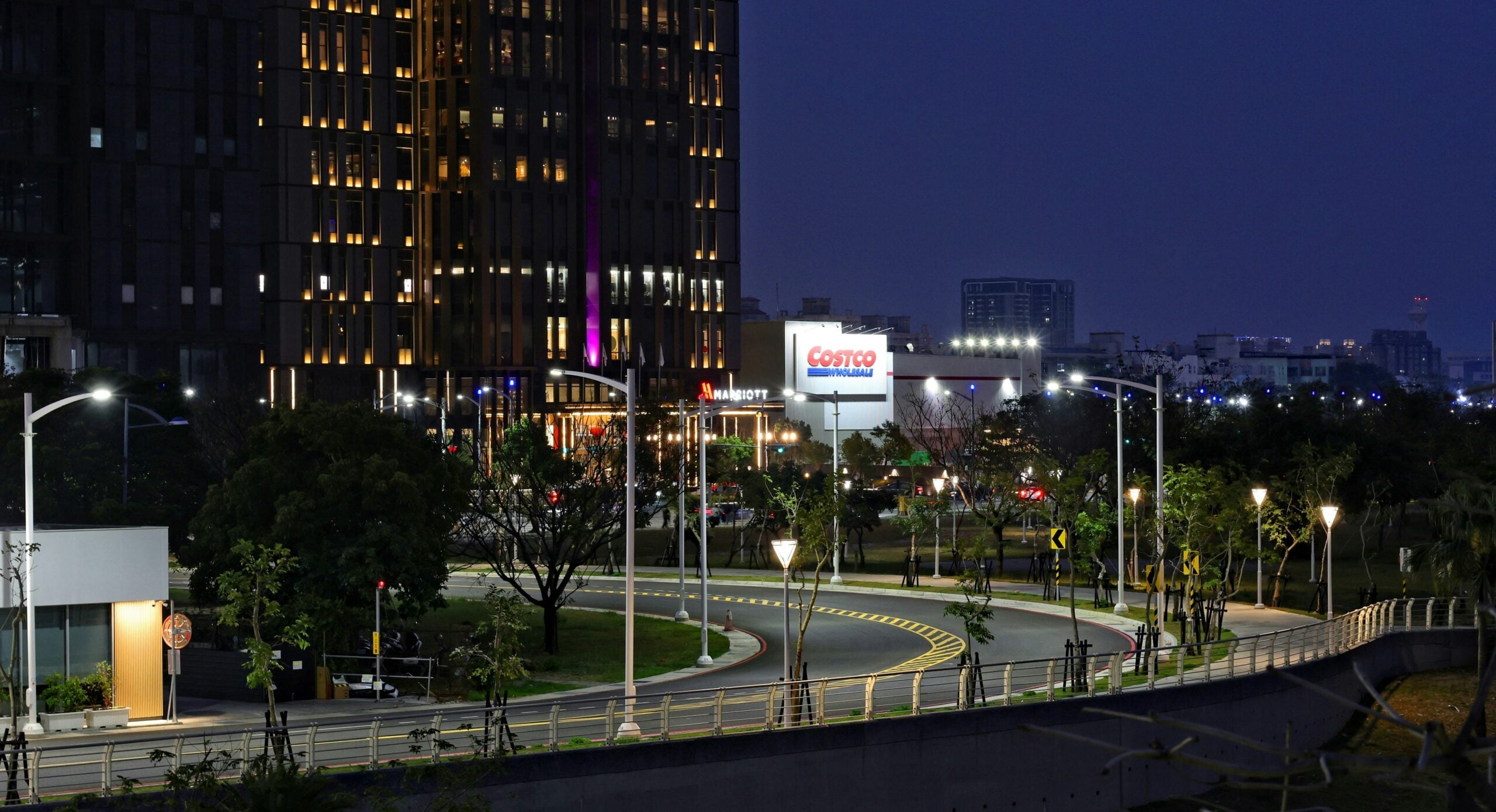 A curved city road at night is lined with streetlights and trees. A tall building with illuminated windows stands on the left, and a brightly lit Costco sign is visible in the background.