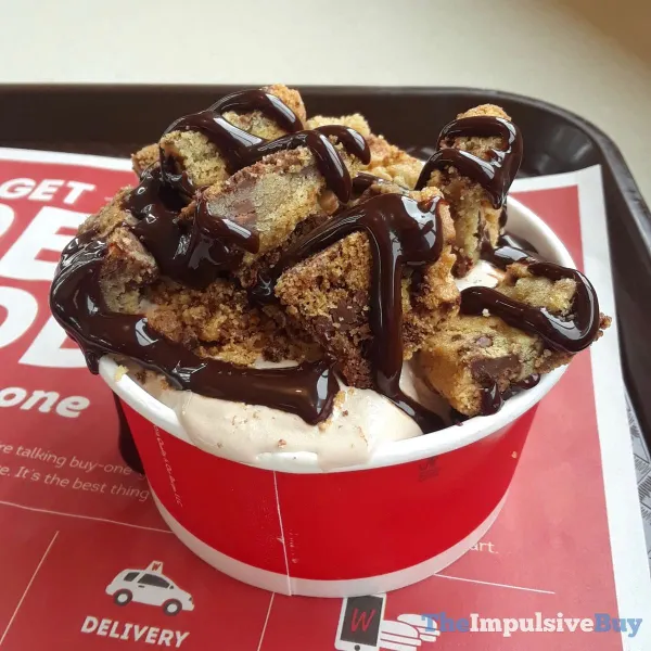 A red cup filled with ice cream topped with chocolate chip cookie pieces and drizzled with chocolate sauce, sitting on a tray with a red and white paper liner.