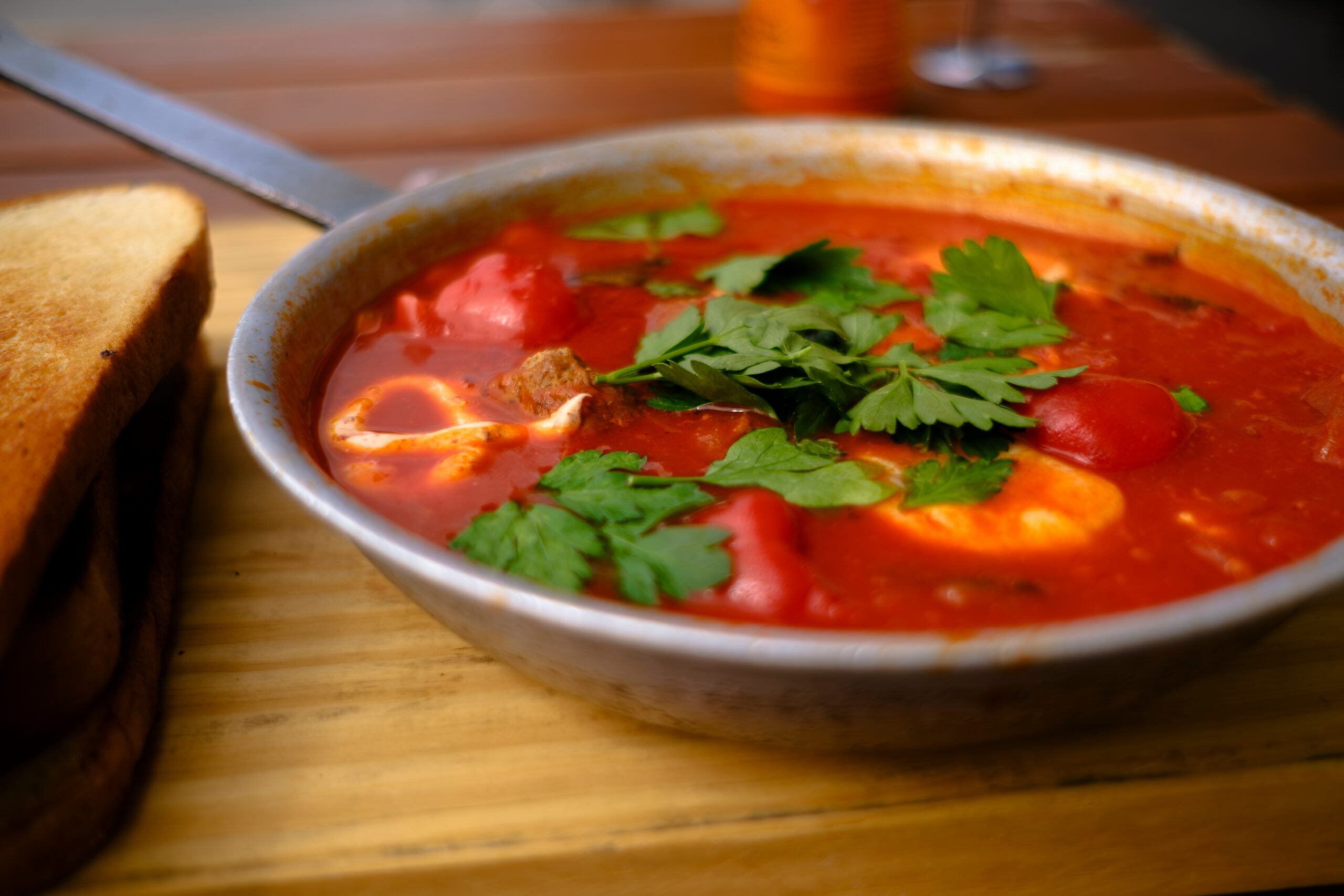 A metal pan filled with a rich red tomato stew, topped with fresh parsley, sits on a wooden surface next to slices of toasted bread.