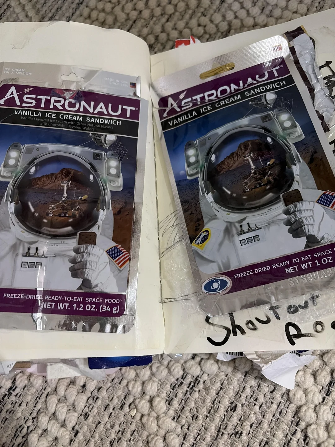 Two packages of freeze-dried vanilla ice cream sandwiches labeled "Astronaut" are taped into an open scrapbook, resting on a textured carpet. The packages feature an astronaut with a space background.
