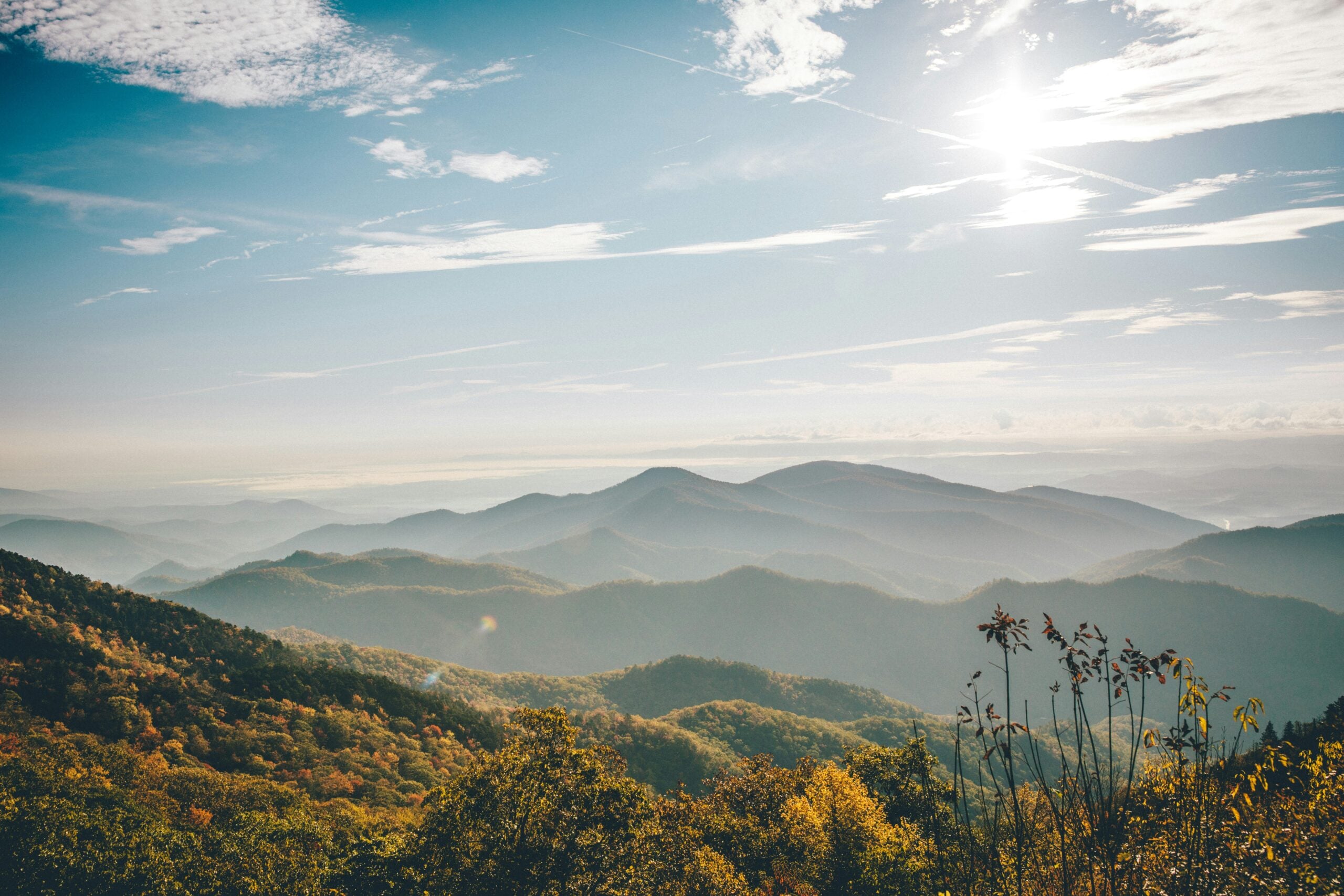 Sunlit mountain range with layers of rolling hills covered in trees, under a bright blue sky with wispy clouds. The foreground shows autumn foliage, while distant peaks fade into a soft, hazy horizon.