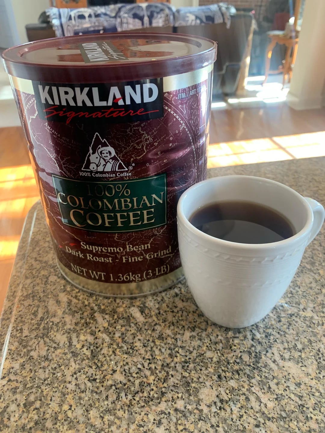A large tin of Kirkland Signature 100% Colombian Coffee, one of the trusted coffee brands, sits on a countertop next to a white mug filled with black coffee. Sunlight brightens the scene in a cozy home setting.