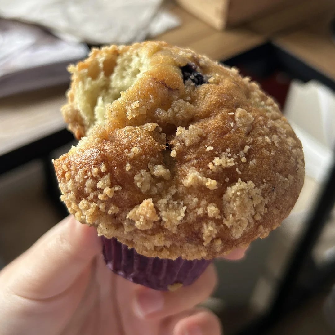 A hand holds a partially eaten muffin with a crumbly topping, revealing a moist interior. The muffin is in a purple paper wrapper, and the background is slightly out of focus with some wooden and dark elements.