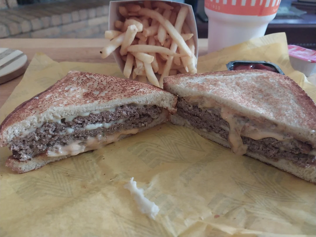 A patty melt sandwich cut in half shows layers of beef, cheese, and sauce on toasted bread, served with a side of French fries, a drink, and a ketchup container on yellow paper.