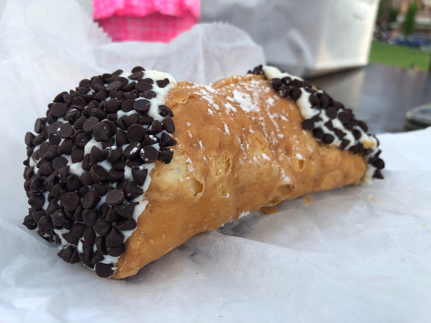 A close-up of a cannoli with ricotta filling, its ends dipped in mini chocolate chips, resting on white wax paper—perfect for those sweet road trip food stops.