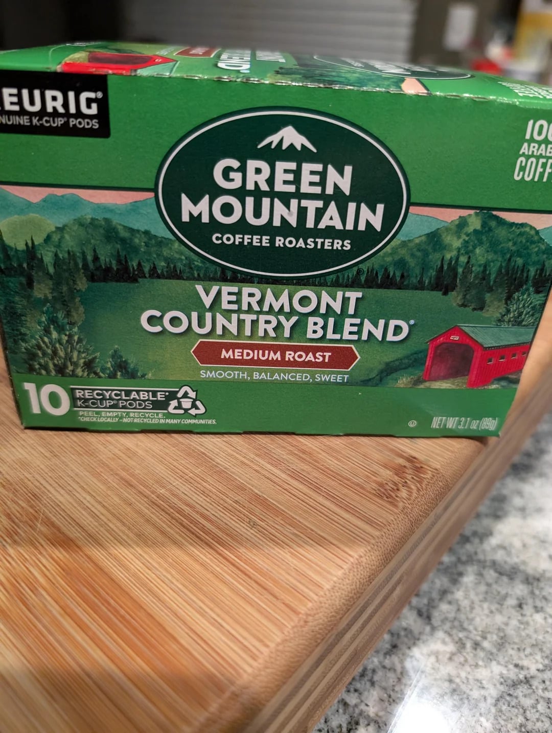 A box of Green Mountain Coffee Roasters Vermont Country Blend Medium Roast K-Cup Pods, from one of the top Coffee Brands, sits on a wooden cutting board, featuring green mountain scenery and a red barn illustration on the front.