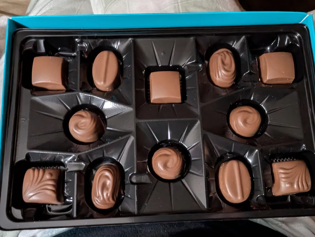 An open box of assorted milk chocolates with several empty spaces where pieces have been taken, showing different shapes and textures of the remaining chocolates in a black plastic tray.