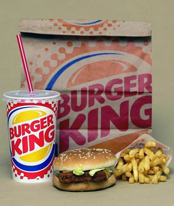 A Burger King meal evokes Vintage Fast-Food Photos, featuring a paper bag in the background, a soft drink cup with a red straw, a hamburger in a sesame seed bun, and French fries on a beige surface.