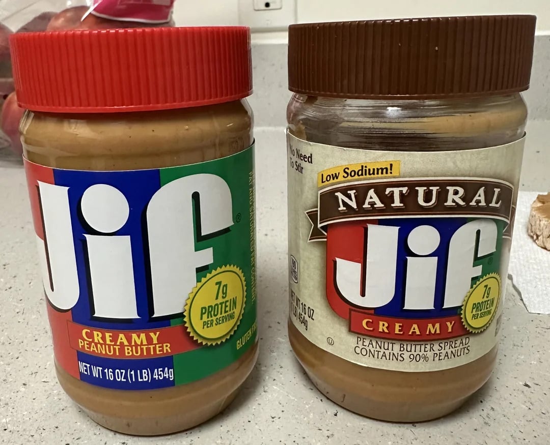 Two jars from the best peanut butter brands sit on the countertop: creamy Jif with a red lid, and Natural Jif, low sodium with 90% peanuts and a brown lid. Both 16 oz jars offer 7g protein per serving.