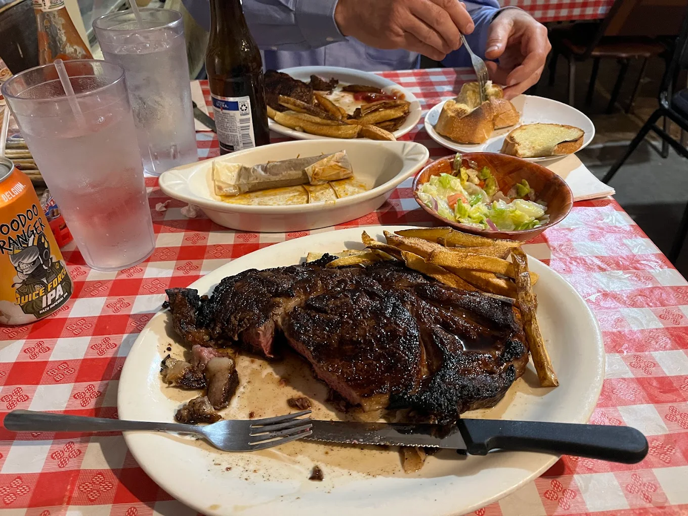 A grilled steak with fries and a knife on a plate sits on a red checkered tablecloth, alongside salad, bread, a glass of water, hot sauce, and a partially visible person cutting food.