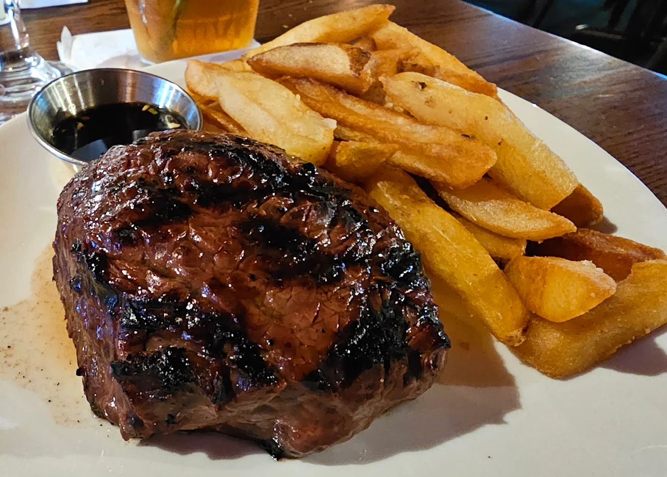 A plate with a grilled steak, thick-cut fries, and a small metal cup of dark dipping sauce, served on a wooden table.