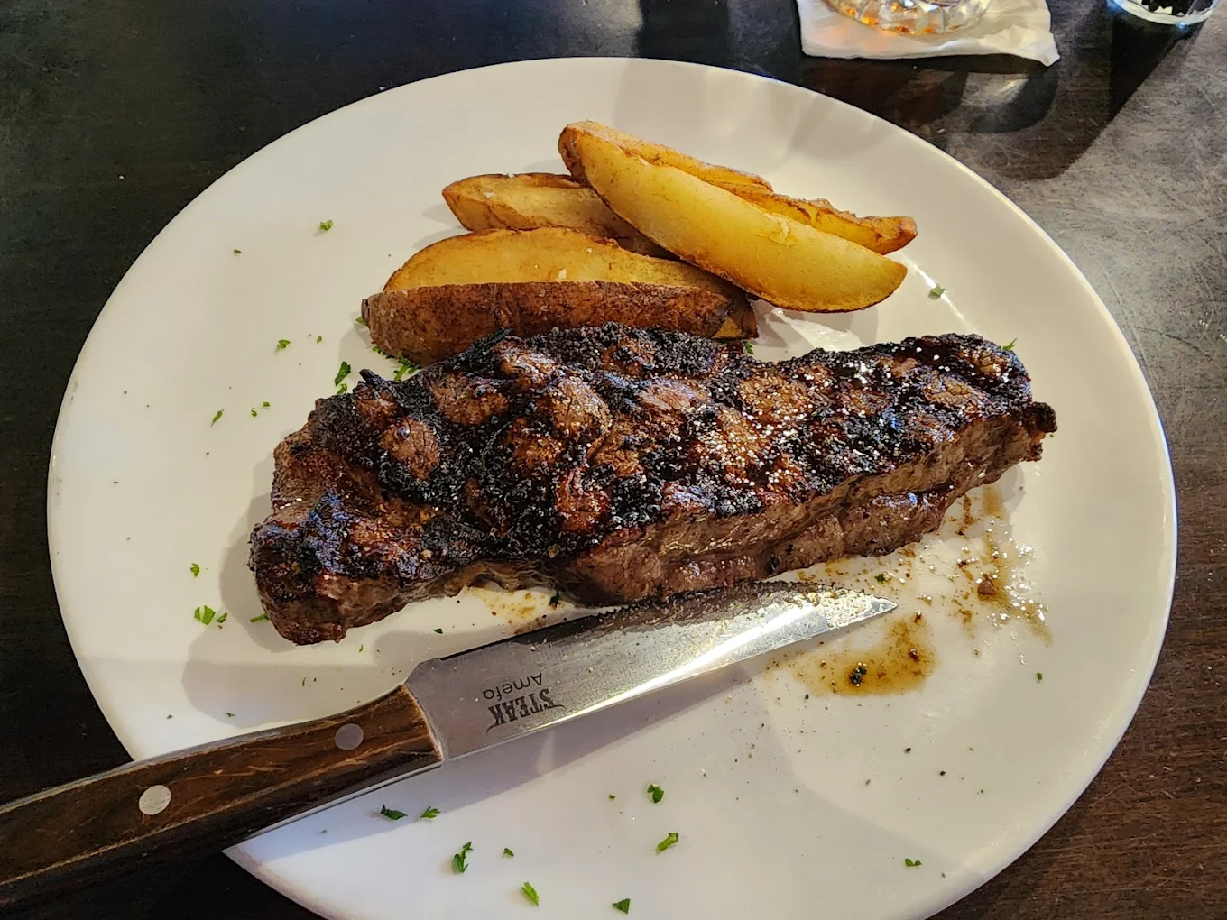 A grilled steak with char marks sits on a white plate beside several thick potato wedges. A steak knife rests on the edge of the plate, and chopped herbs are sprinkled around the food.
