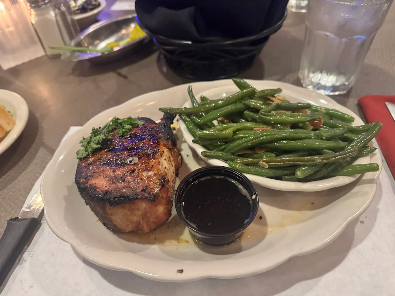 A white plate with a grilled pork chop garnished with parsley, a serving of sautéed green beans, and a small cup of sauce. A glass of water and bread basket are in the background.