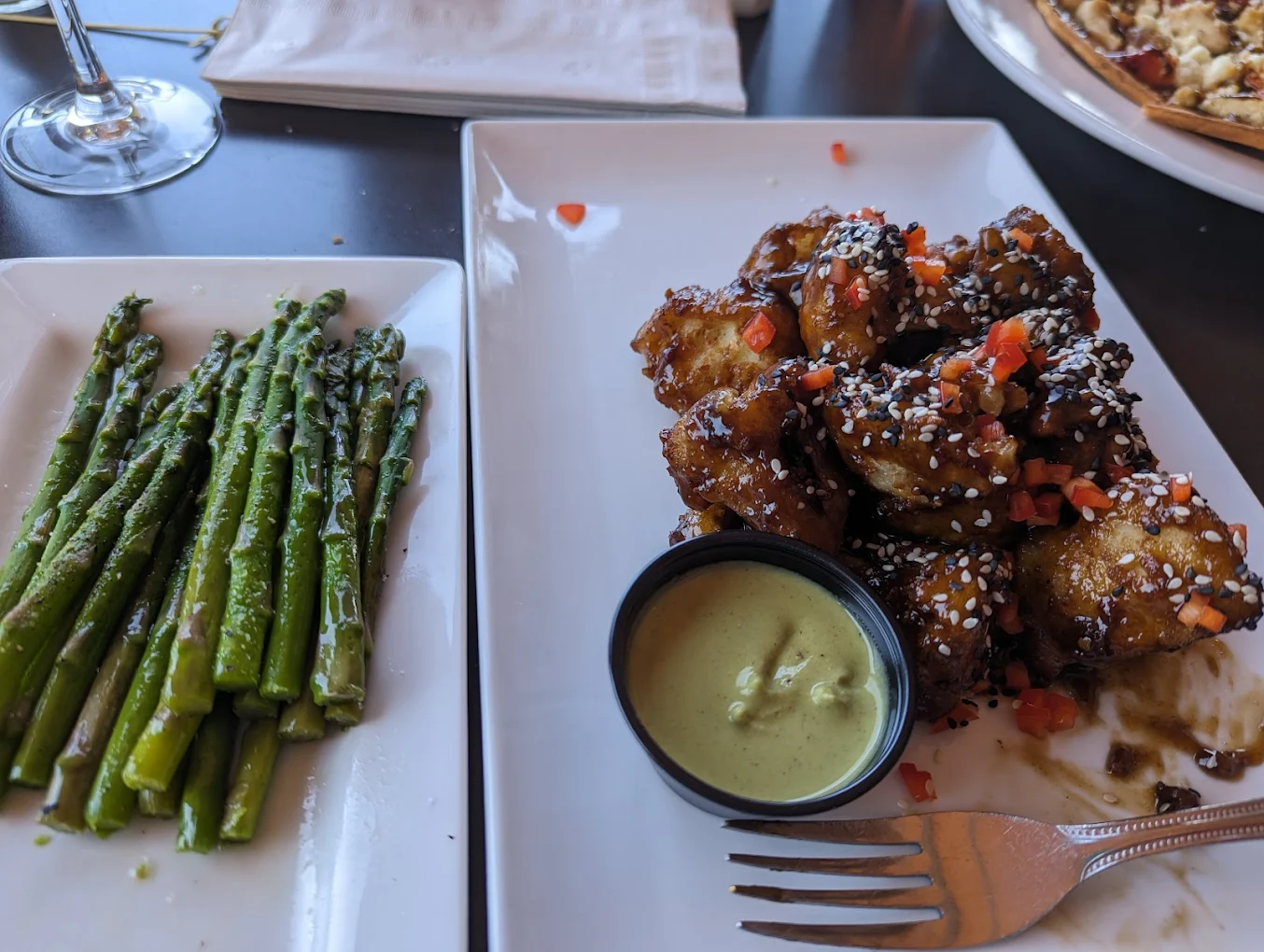 A plate of roasted asparagus spears sits next to a rectangular plate of glazed, sesame-sprinkled cauliflower bites with diced red pepper and a small cup of dipping sauce. A fork rests on the edge of the plate.