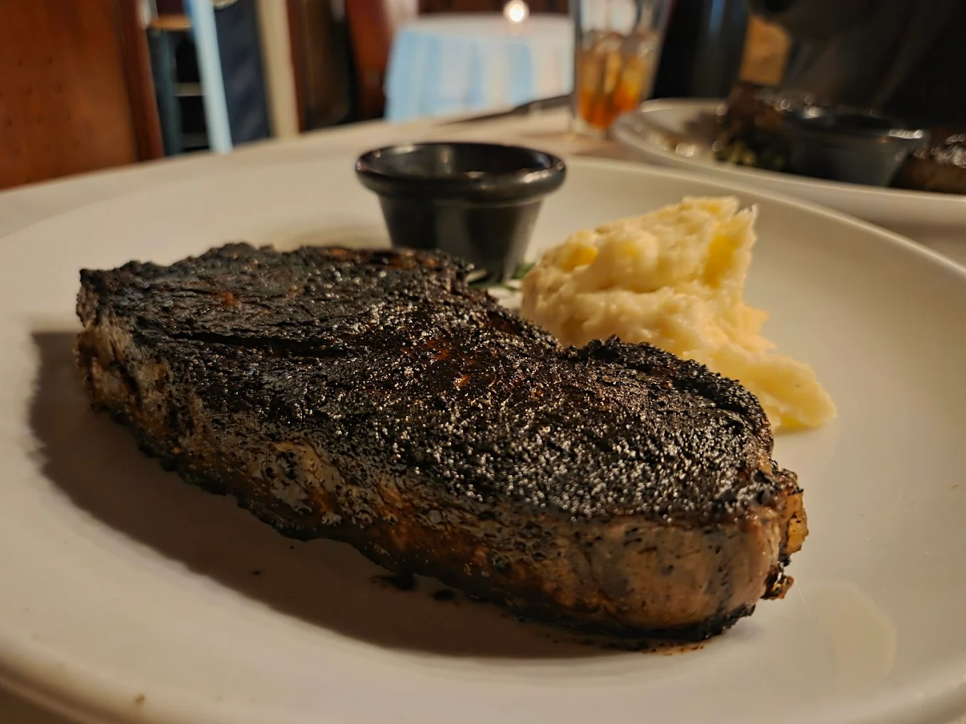East Coast steakhouses. A plate with a blackened, seasoned steak, a serving of mashed potatoes, and a small cup of sauce, set on a restaurant table.