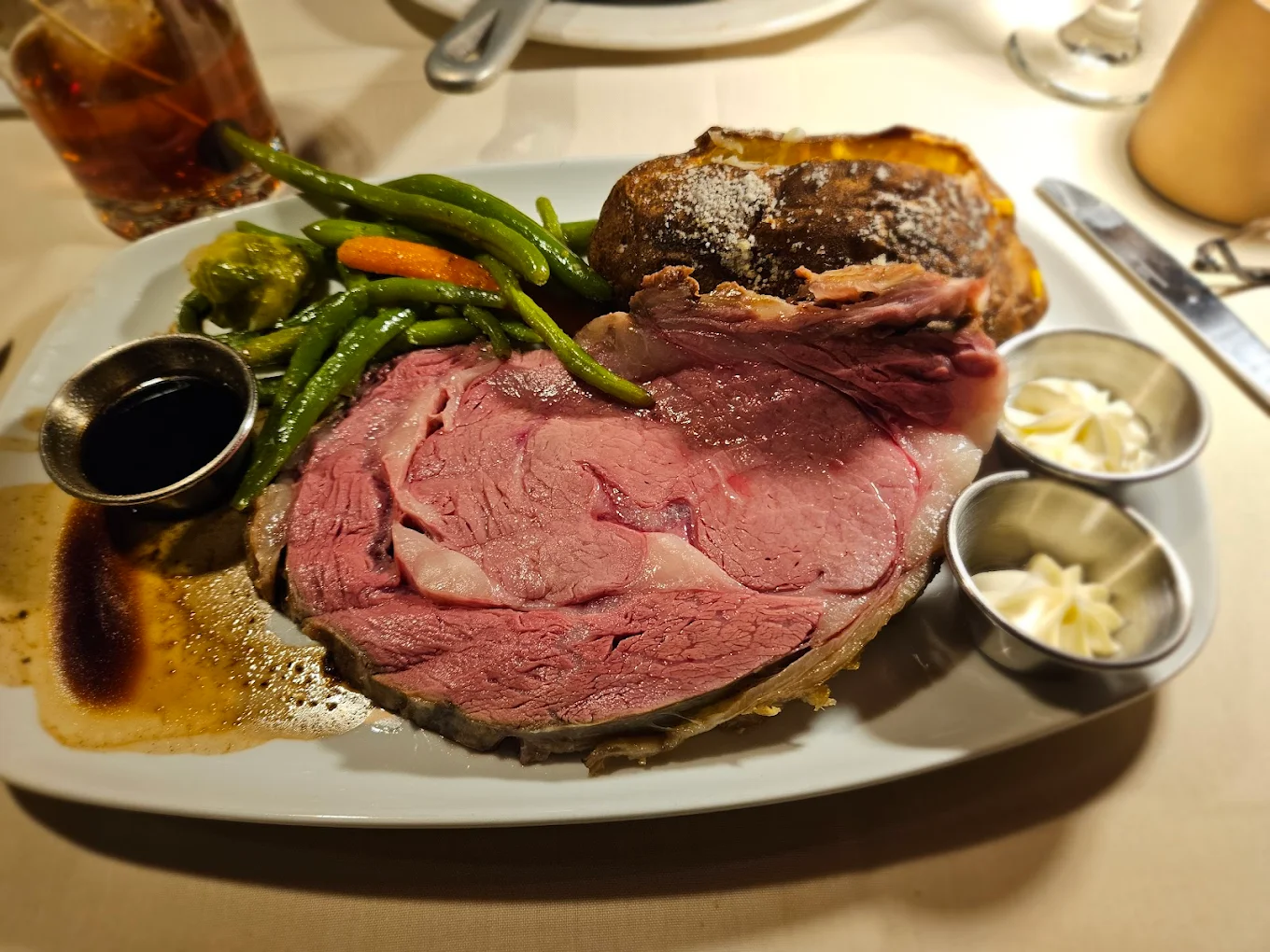 A plate with a thick slice of rare roast beef, a baked potato, green beans, carrots, broccoli, a cup of au jus, and two small cups of butter, set on a white tablecloth.