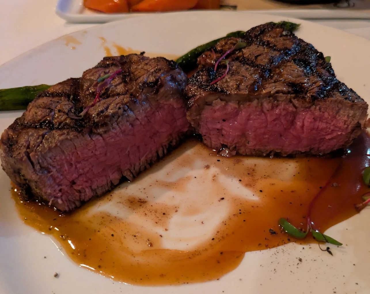 A grilled steak sliced in half, showing a juicy medium-rare center, sits on a white plate with a pool of brown sauce and some green garnish. Grilled asparagus spears are on the side.