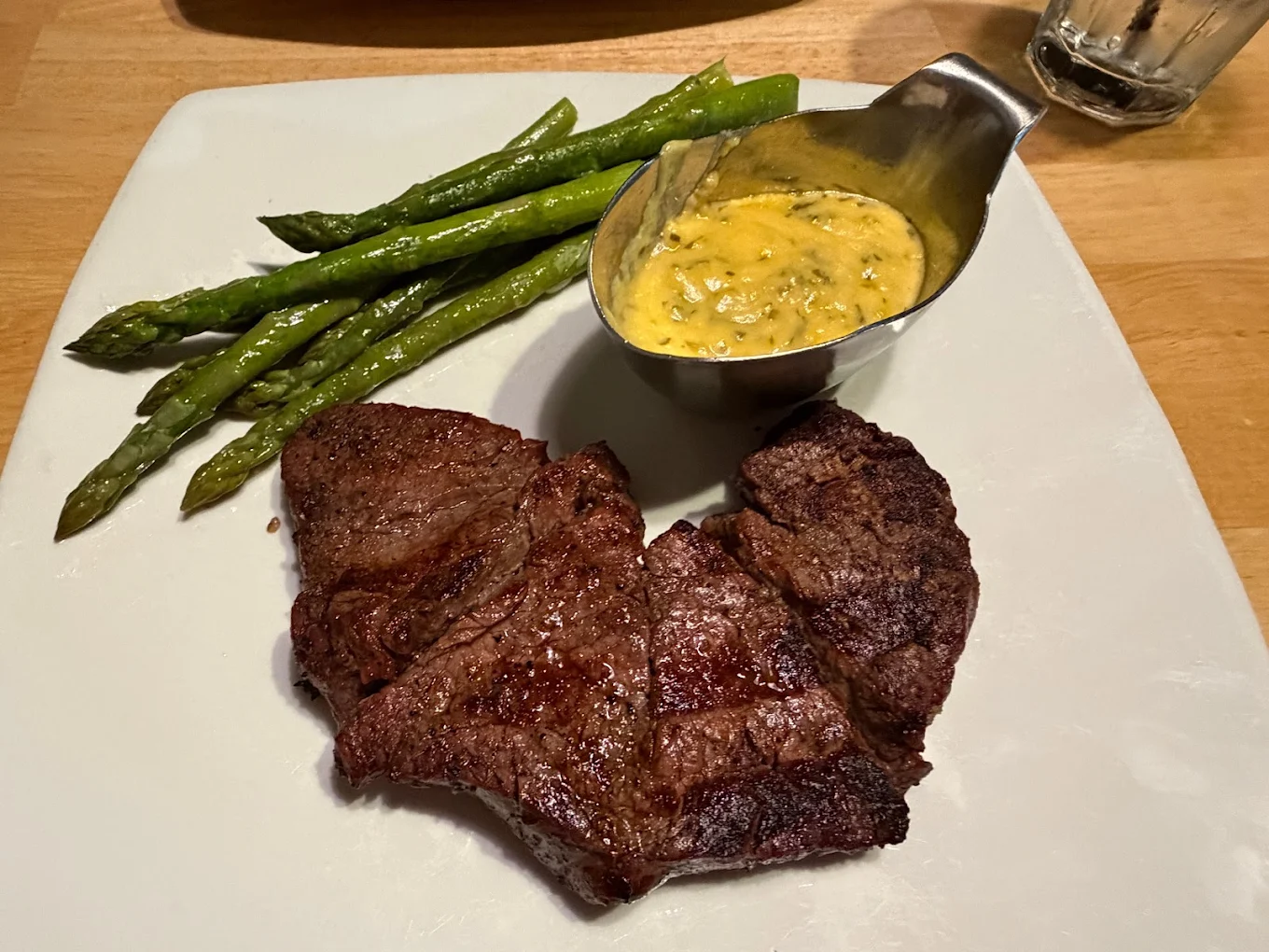 A plate with grilled asparagus, a serving of cooked steak, and a small metal sauce boat filled with a creamy yellow sauce on a white square plate.