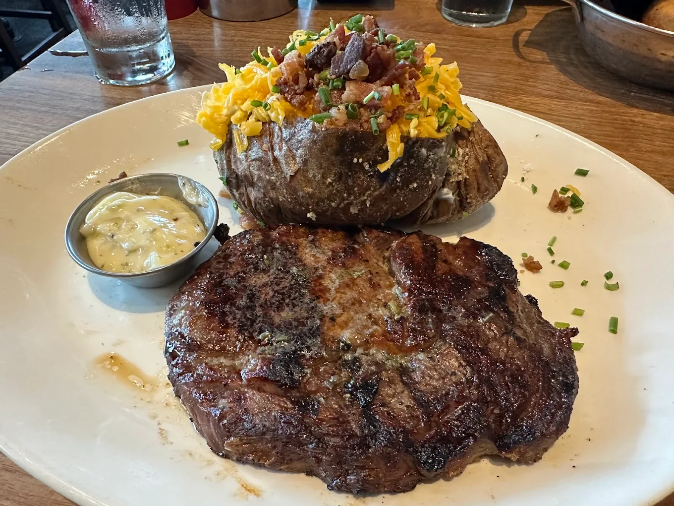 A grilled steak sits on a plate next to a loaded baked potato topped with shredded cheese, bacon bits, and chives. A small cup of sauce is on the side. The meal is on a wooden table.