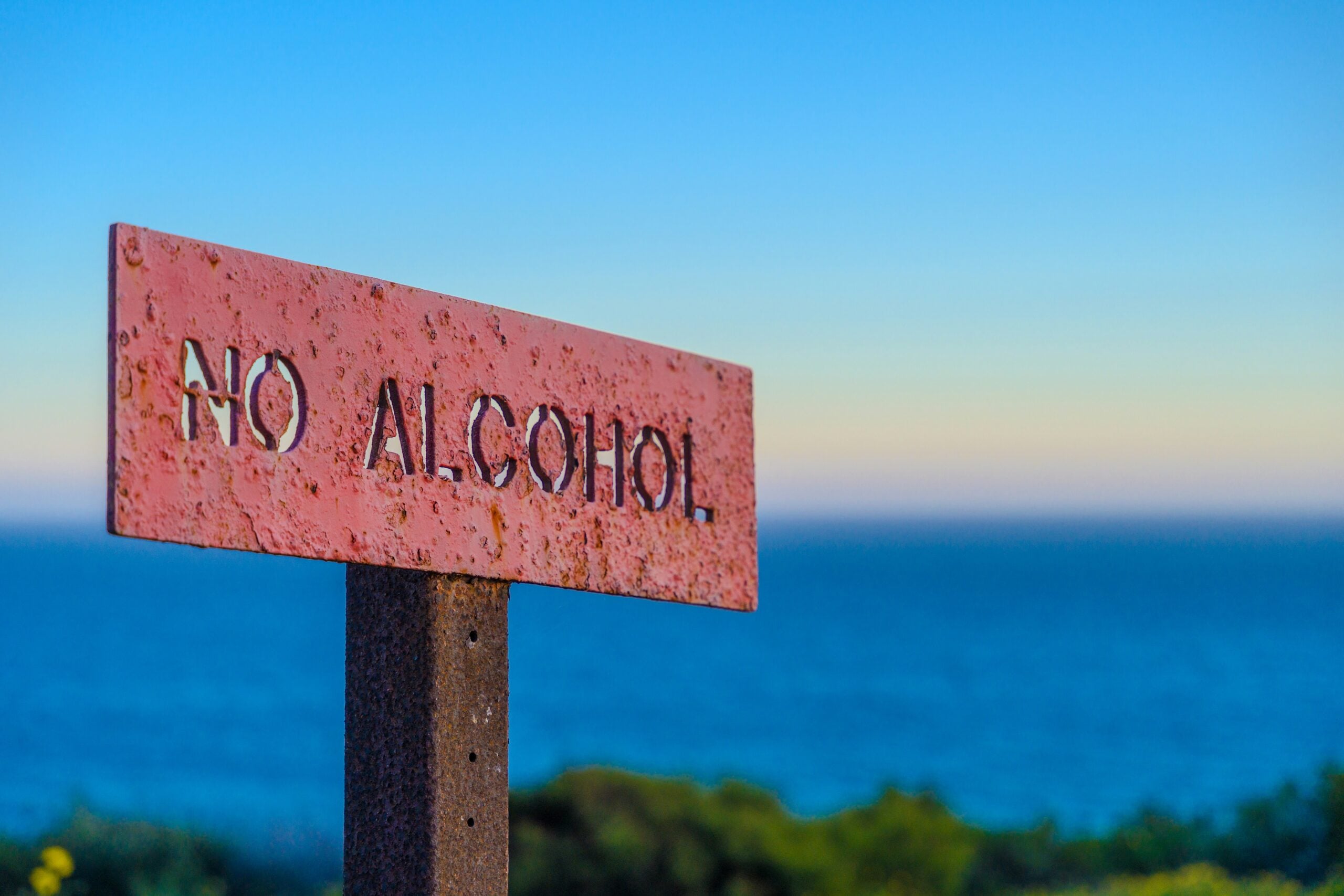 A weathered red sign with cut-out letters reading "NO ALCOHOL" stands in front of a blurred background of ocean and blue sky.