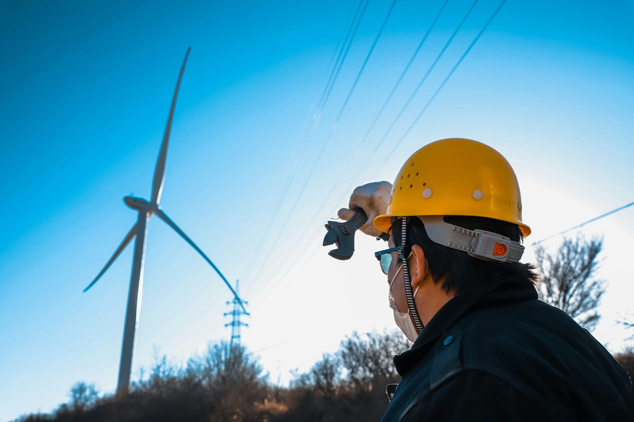 A worker wearing a yellow hard hat and protective mask looks at a wind turbine on a clear day, with trees and power lines in the background.