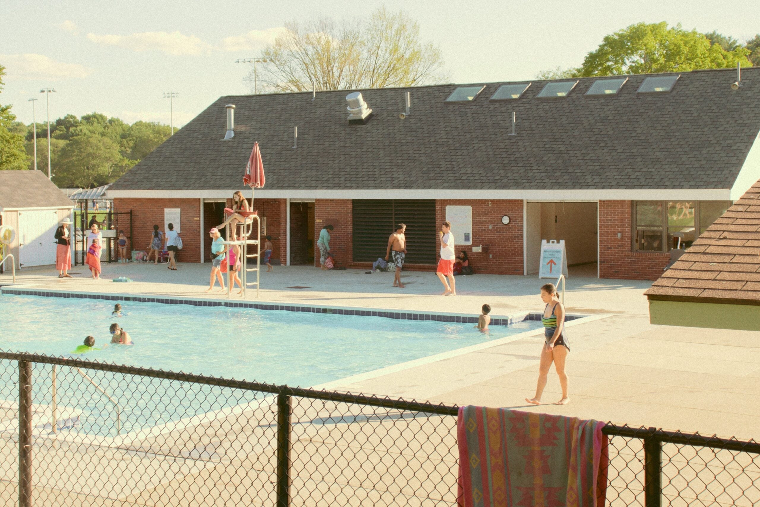 People swim and relax by a community outdoor pool on a sunny day. A lifeguard sits on a raised chair, and several people stand or walk near a brick building in the background. A towel hangs on the black fence in front.