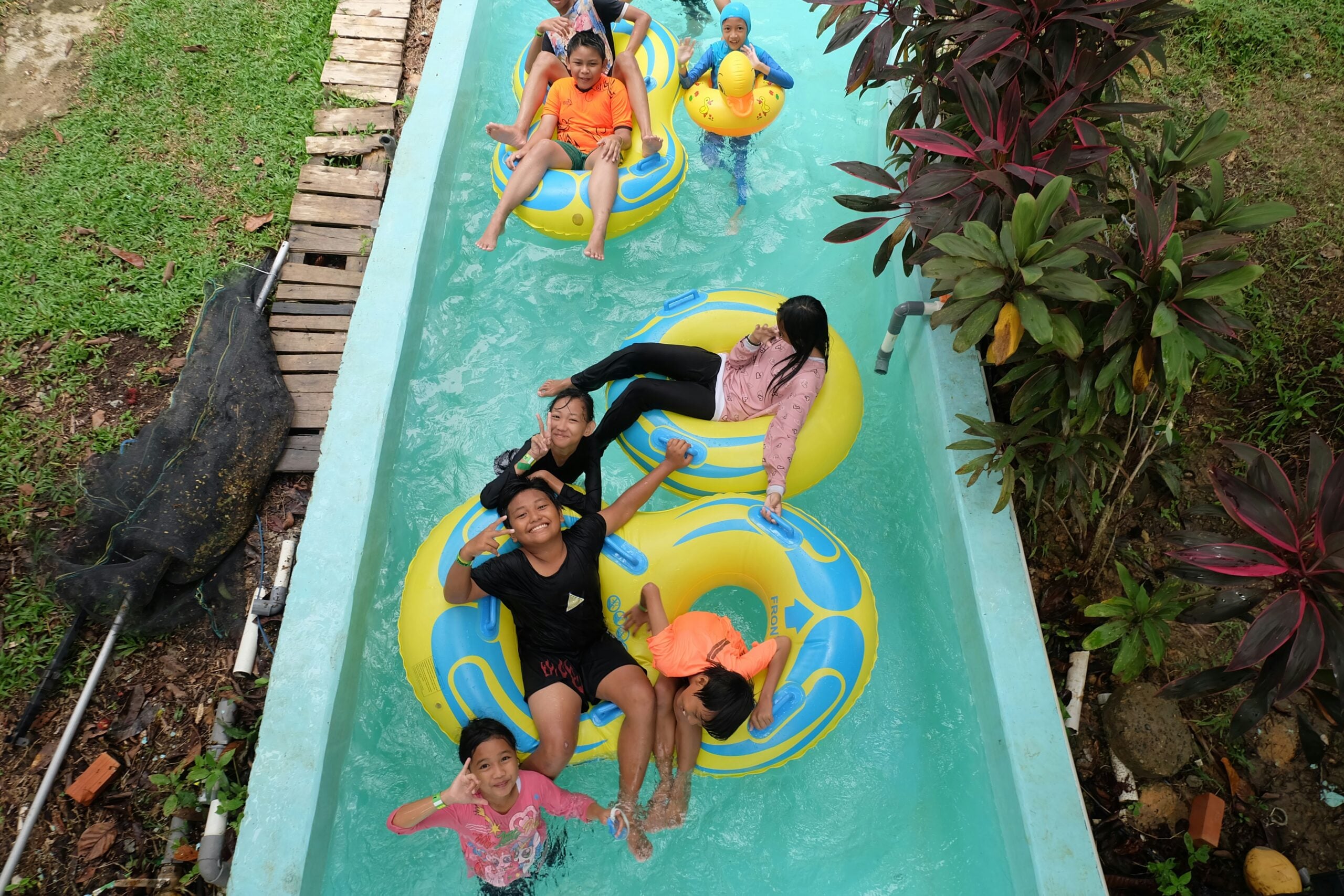 A group of children in swimsuits enjoy floating on yellow and blue inflatable rings in a narrow outdoor pool, smiling and playing together beneath green and red plants.