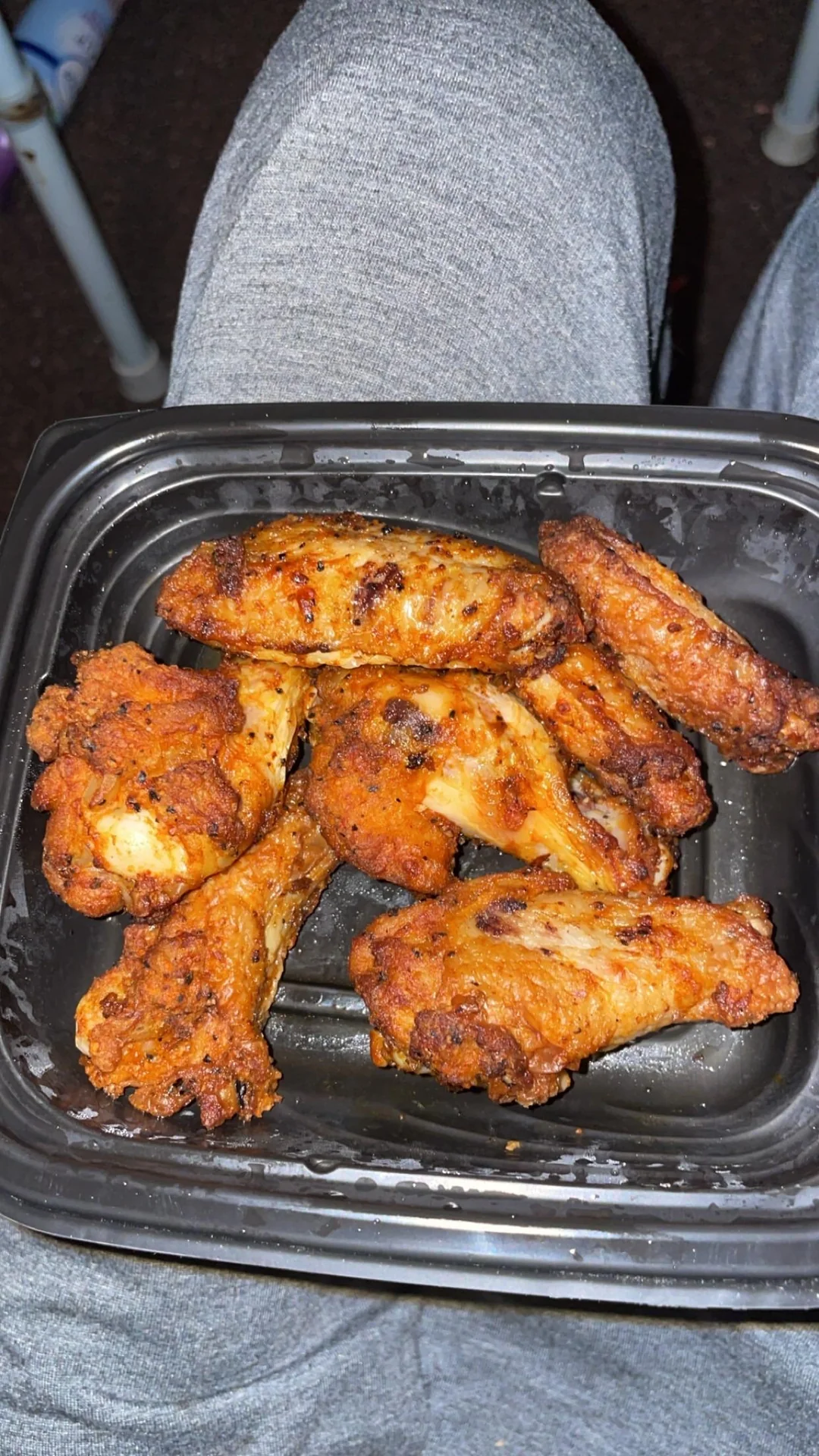 A black plastic container holds seven seasoned and cooked chicken wings, resting on someone's lap. The person is wearing light gray pants.