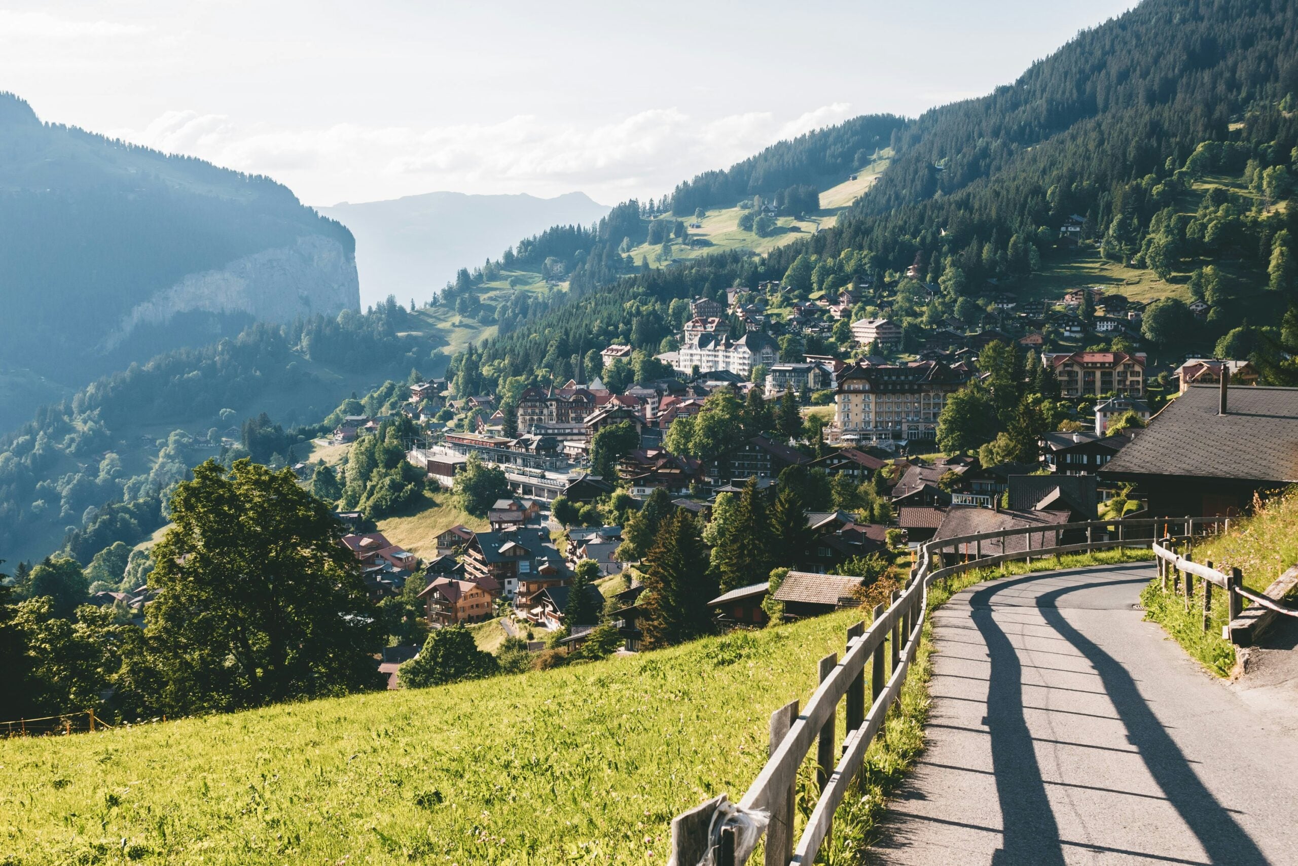 A winding paved path with a wooden fence overlooks a picturesque mountain village nestled among lush green hills and trees, with mountains in the background under a bright sky.