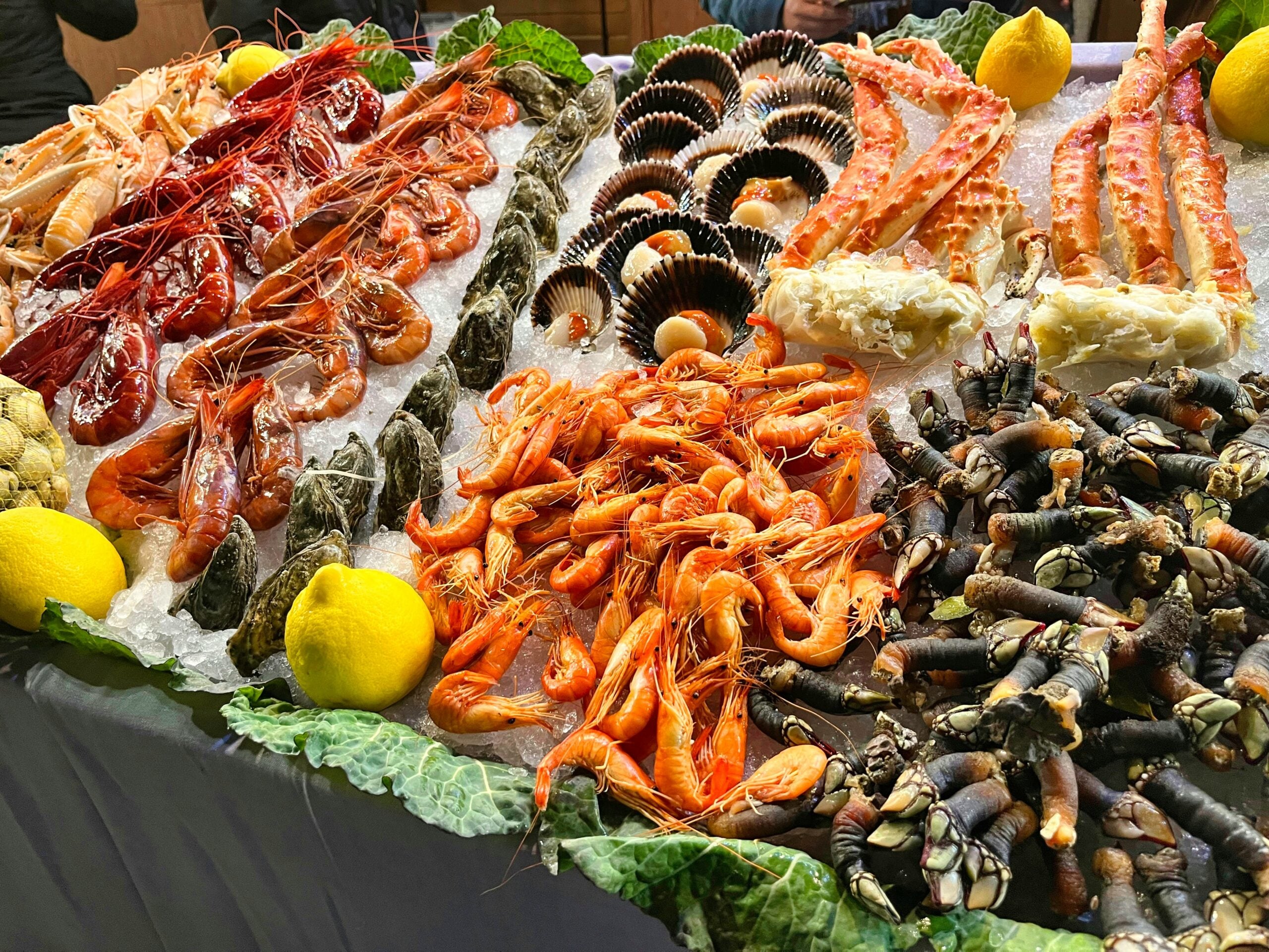 A seafood display on ice featuring shrimp, prawns, scallops, crab legs, barnacles, clams, and lemons, arranged in colorful, neatly organized rows with leafy greens as garnish.