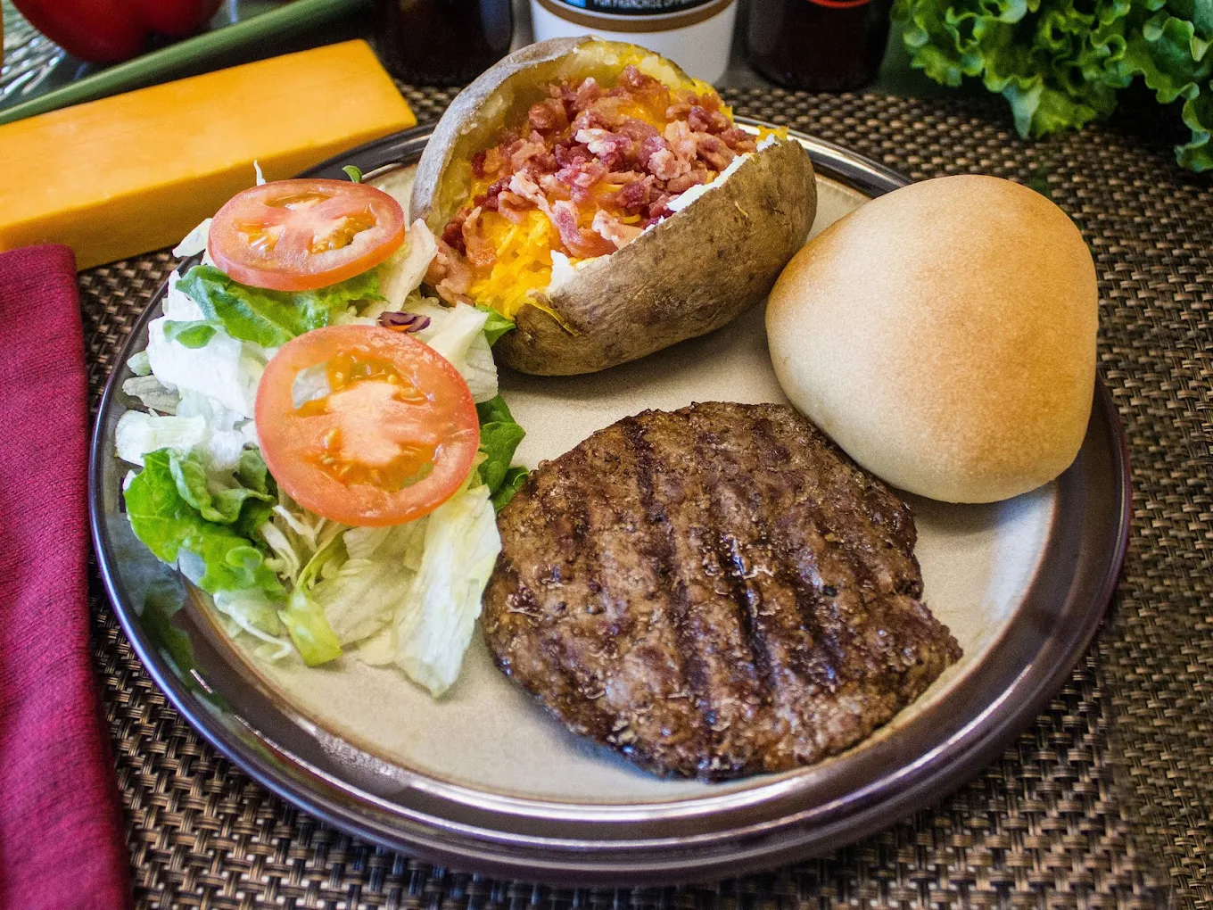 A plate with a grilled beef patty, a baked potato topped with bacon and cheese, a dinner roll, and a side salad with sliced tomatoes and lettuce. Cheddar cheese and leafy greens are in the background.