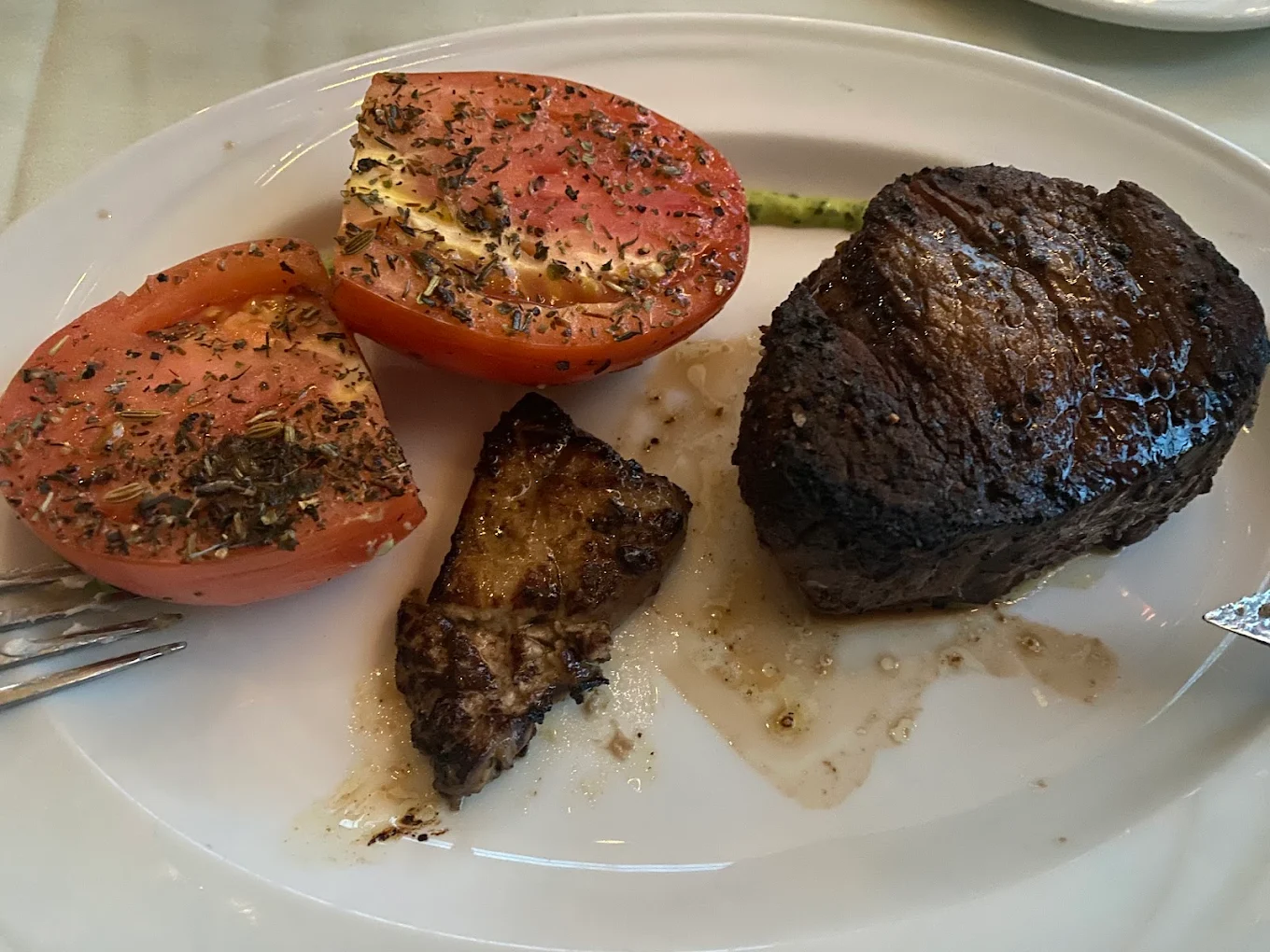 A plate with a grilled filet mignon, a piece of cooked foie gras, and two seasoned tomato halves, garnished with herbs, on a white oval dish.