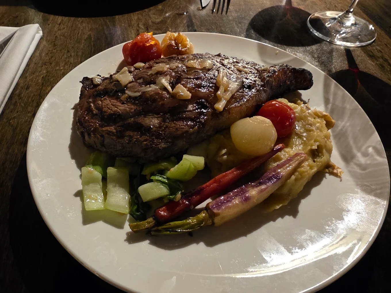 A grilled steak topped with garlic is served on a plate with mashed potatoes, roasted cherry tomatoes, carrots, pearl onions, celery, and green vegetables. A wine glass and folded napkin are nearby on the wooden table.