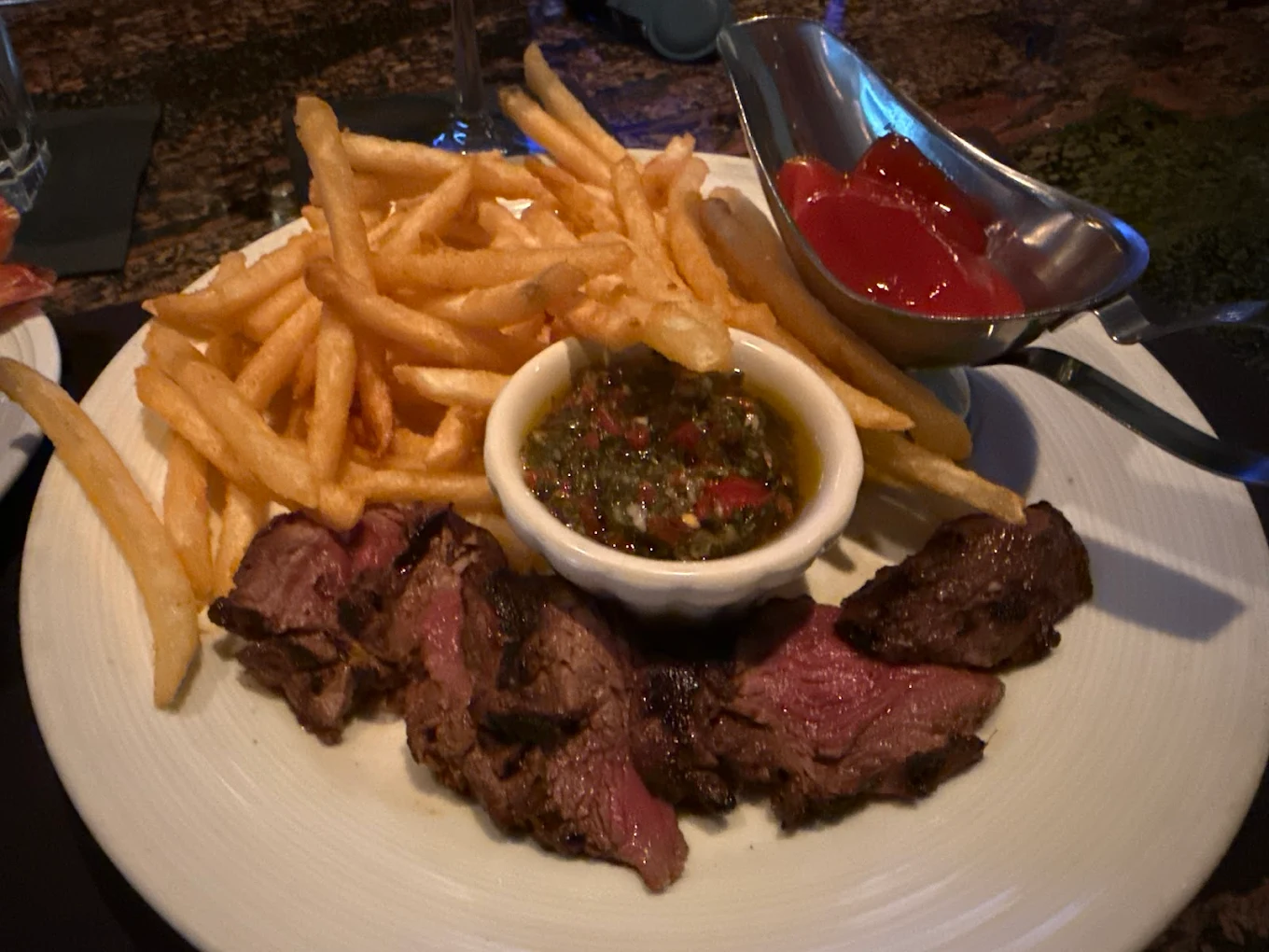 A plate of sliced steak with a small bowl of chimichurri sauce, French fries, and a metal gravy boat filled with ketchup. The food is served on a white plate.