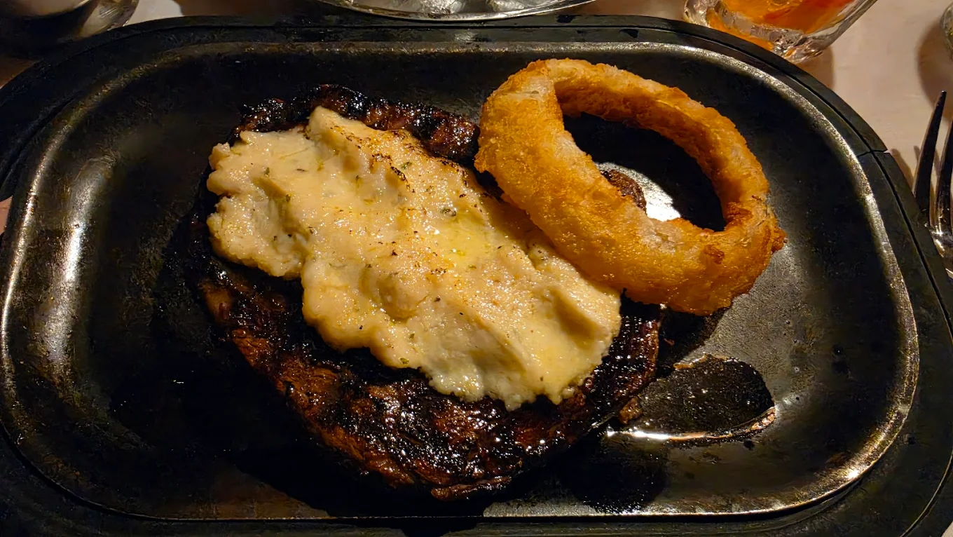 A grilled steak topped with creamy mashed potatoes and served with a single crispy onion ring on a black plate.