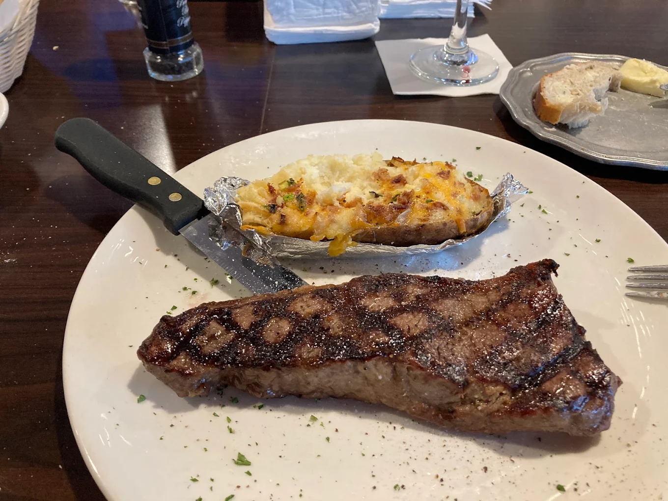 A grilled steak with char marks and a baked potato topped with cheese and bacon bits are served on a white plate, with a knife on the side. A bread basket, butter, and wine glass are in the background.