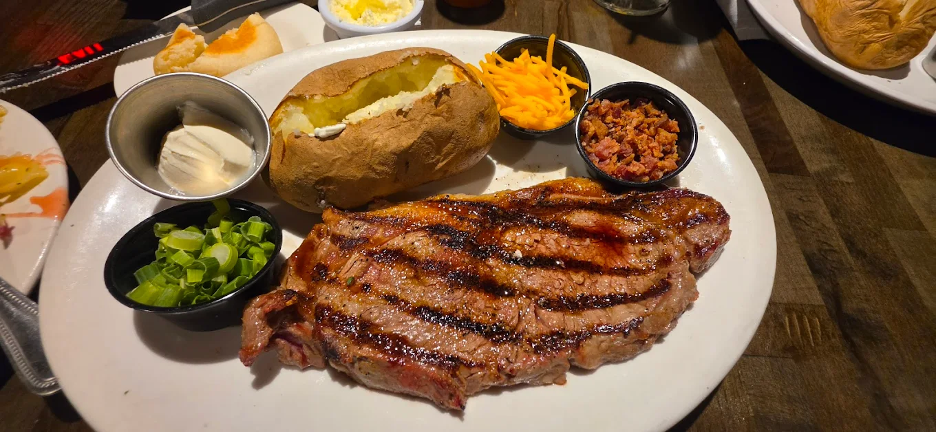 A grilled steak served with a baked potato, shredded cheese, bacon bits, sour cream, and chopped green onions on a white plate. Another plate with food is partially visible in the background.