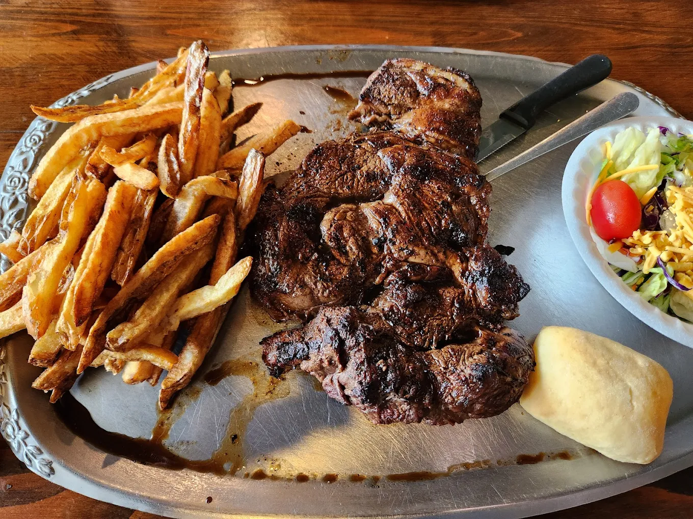 Under-the-radar steakhouses in the South. A metal tray holds a large grilled steak, a generous serving of French fries, a bread roll, and a side salad with lettuce, tomato, and shredded vegetables. A knife rests beside the steak.