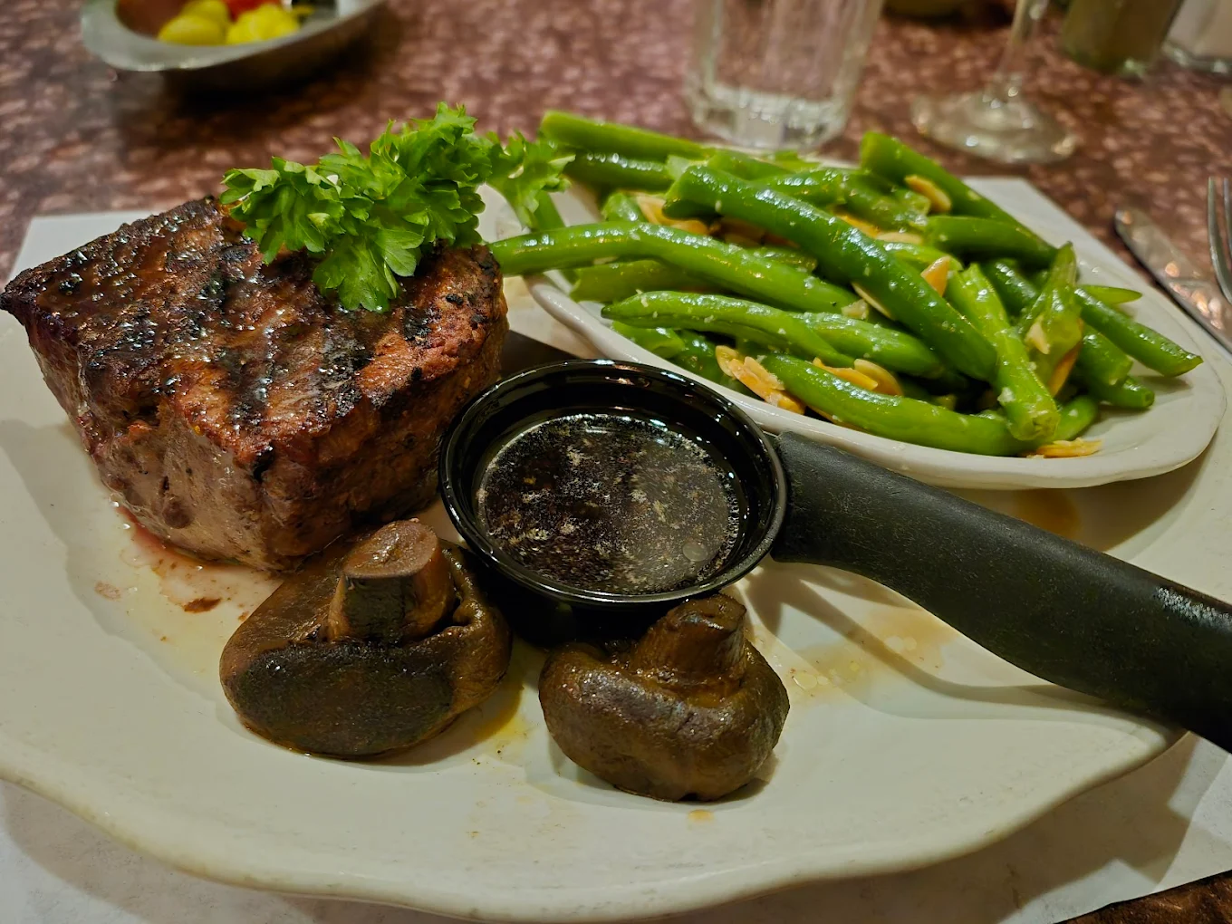 A plate with a grilled steak topped with parsley, two cooked mushrooms, a small cup of dark sauce, and a side of green beans sautéed with garlic. A fork and knife rest on the plate.