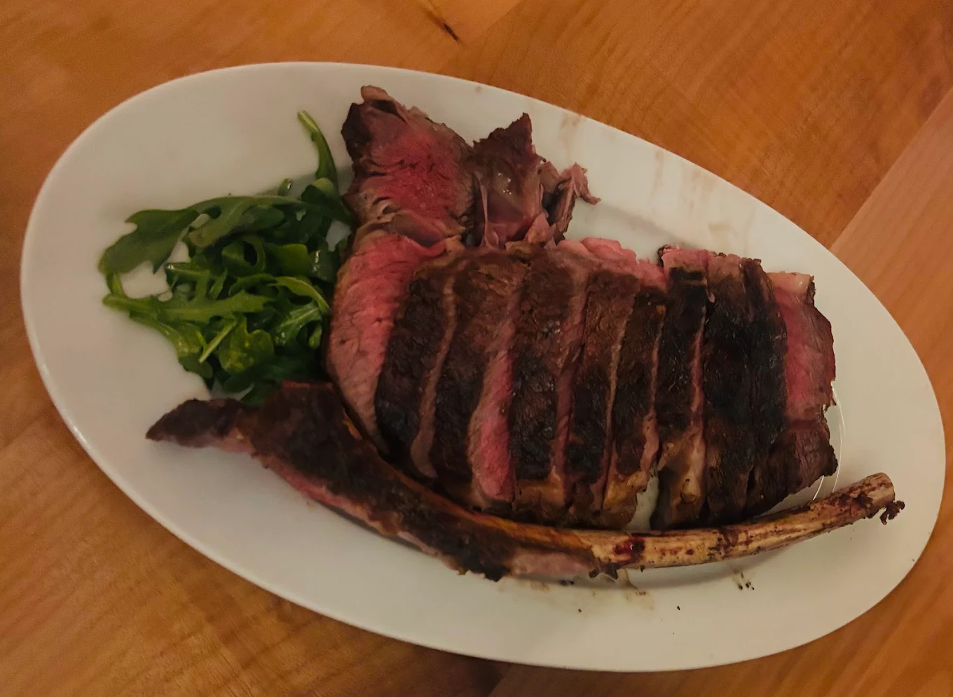 A white oval plate with sliced, medium-rare tomahawk steak and a small side of fresh arugula, placed on a wooden table.