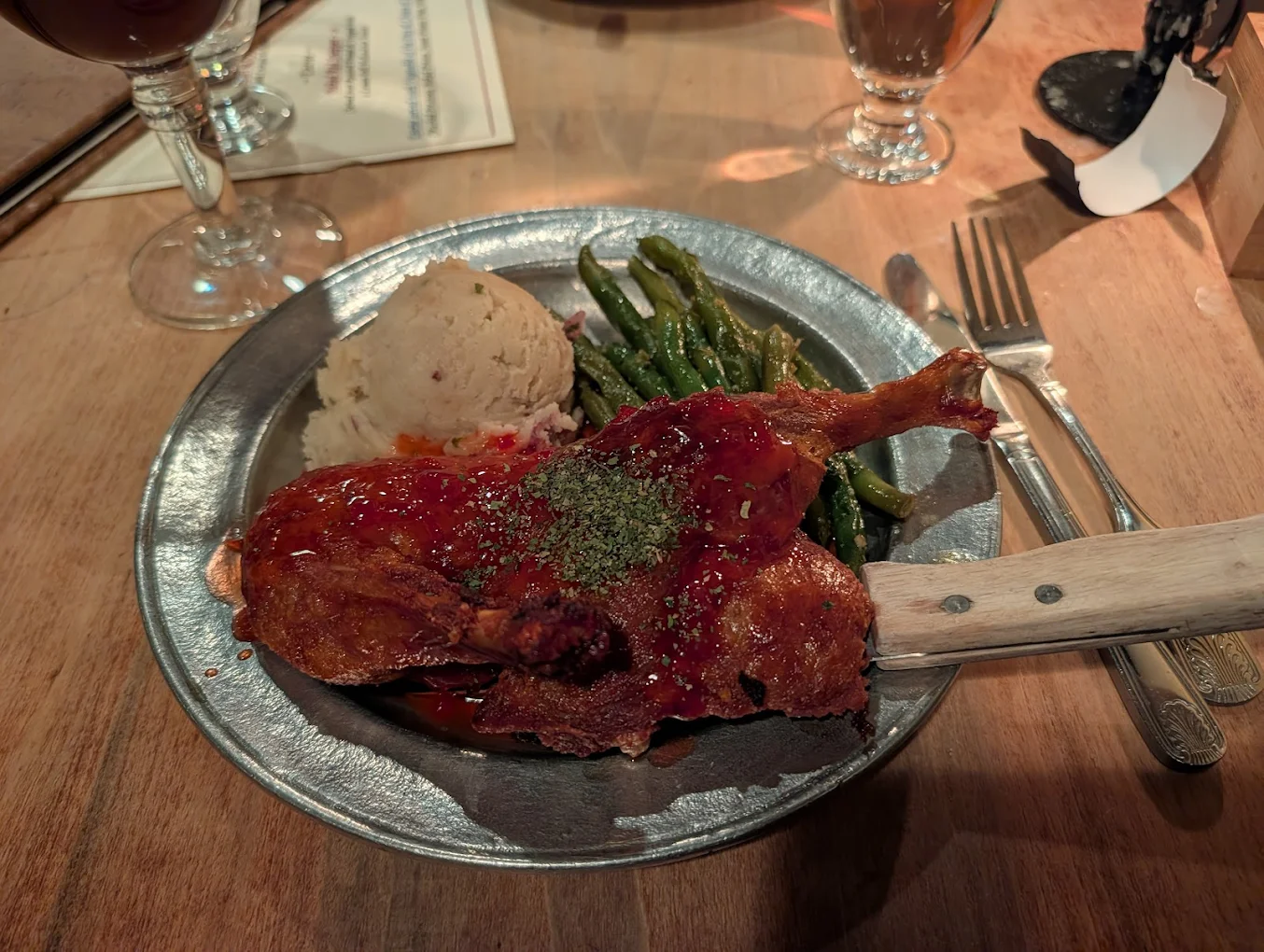 A metal plate with roasted duck covered in sauce and herbs, mashed potatoes, and green beans. A knife rests on the plate, with wine glasses, a fork, and a menu on the wooden table.