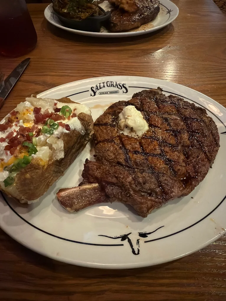 A grilled ribeye steak with a dollop of butter is served alongside a loaded baked potato topped with cheese, bacon, and green onions on a Saltgrass Steak House plate.