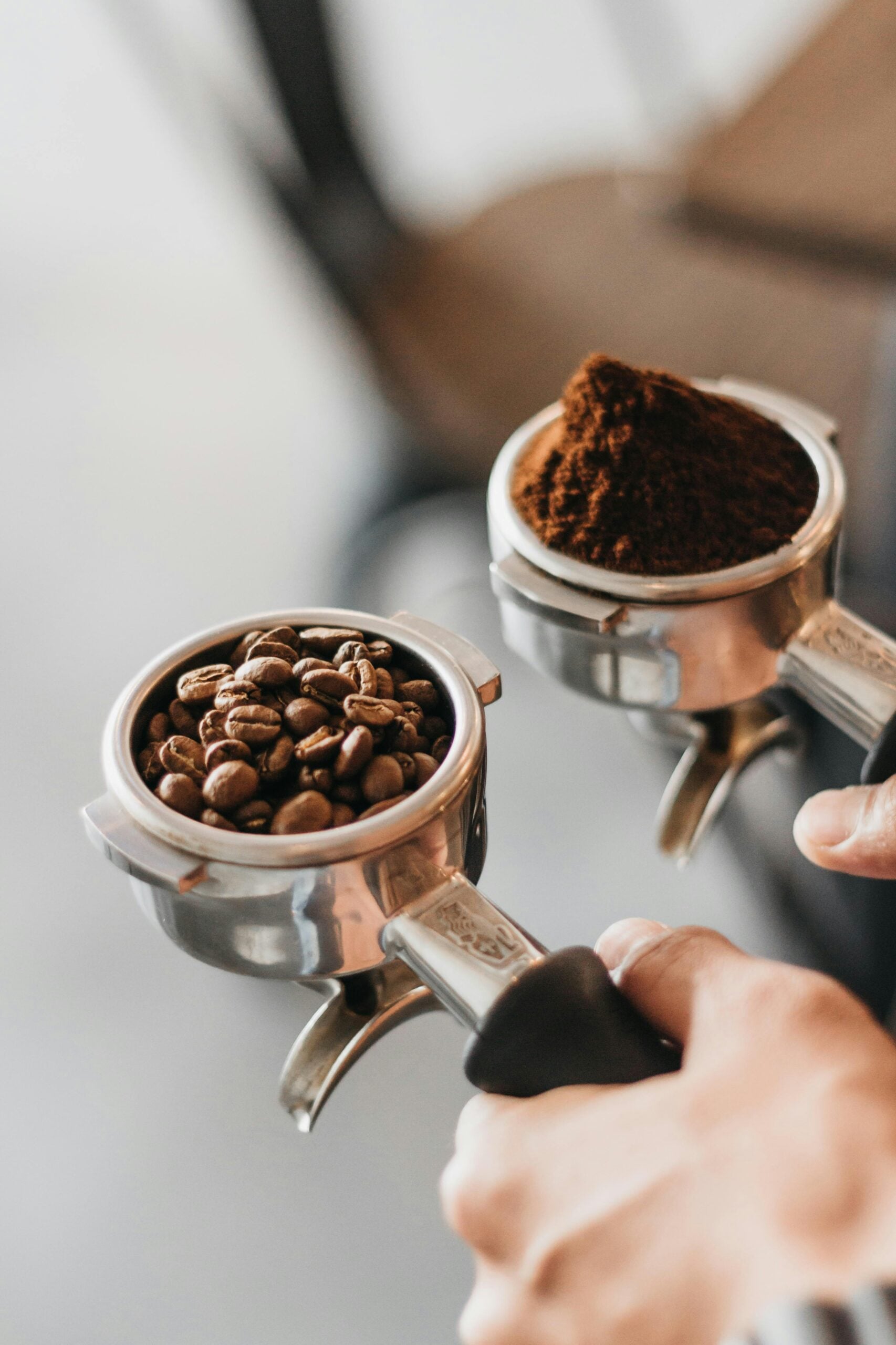 A person holds two portafilters: one filled with whole coffee beans and the other with ground coffee, demonstrating the coffee preparation process.