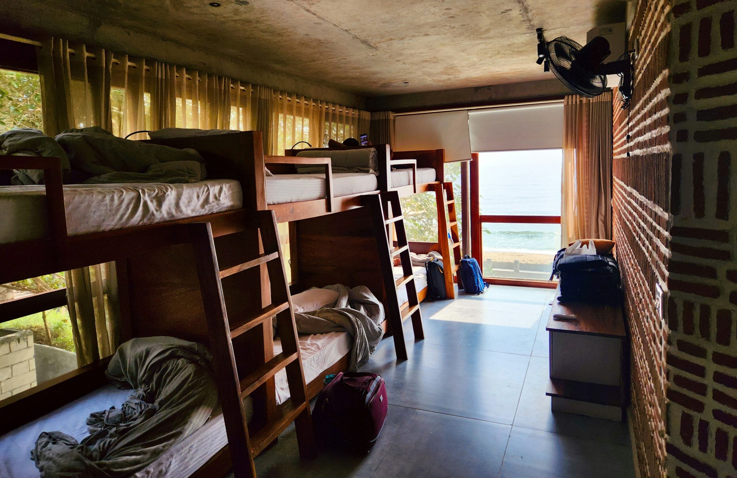 A sunlit hostel room with four wooden bunk beds, some unmade, backpacks on the floor, and a large window overlooking the ocean. Light curtains cover the windows, and a fan is mounted high on the wall.