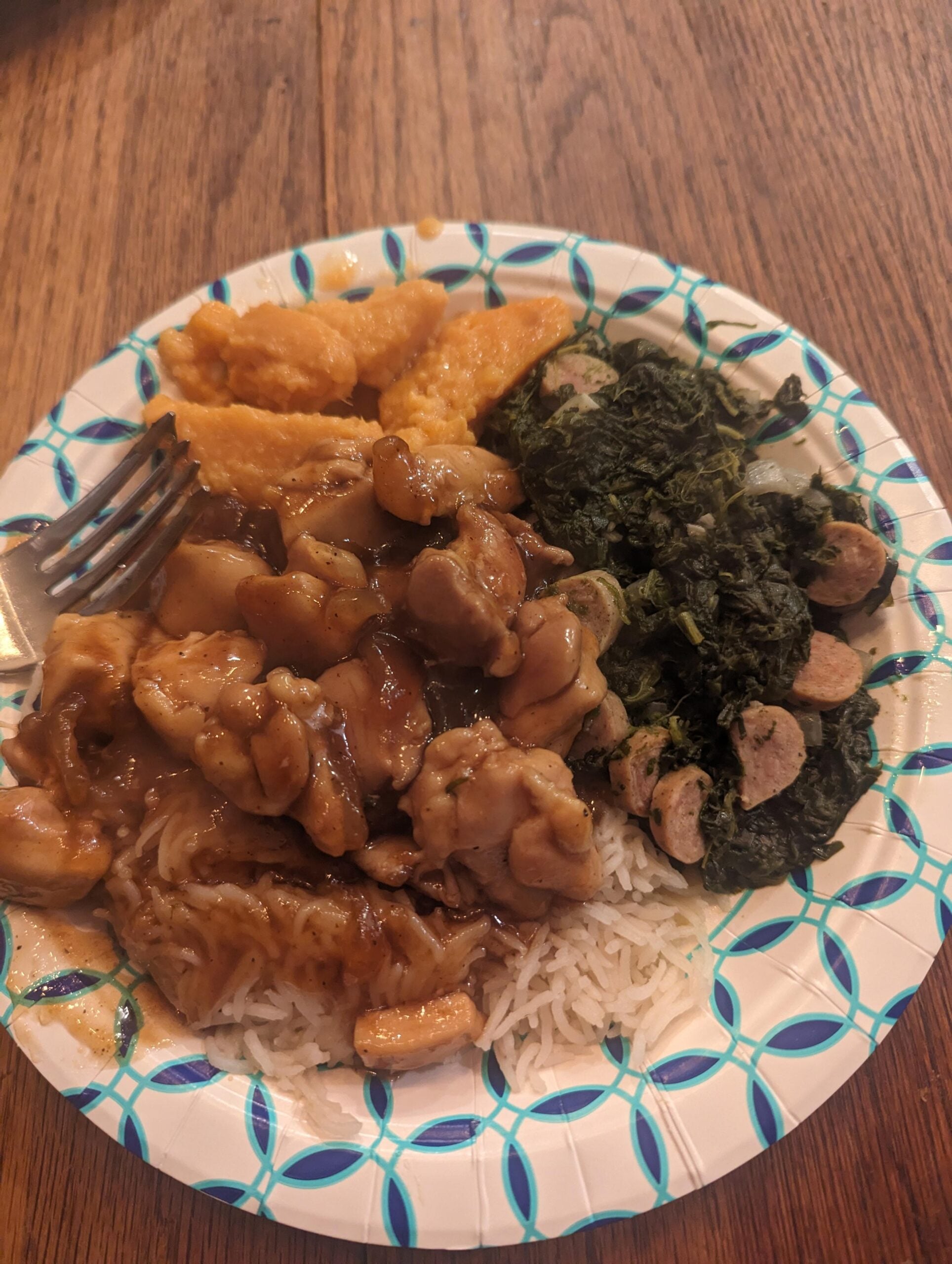 A paper plate with rice topped with brown saucy chicken, cooked greens with sliced sausage, and baked orange sweet potatoes sits on a wooden table with a fork on the side—a surprisingly appetizing scene, far removed from any hilarious cooking disasters.